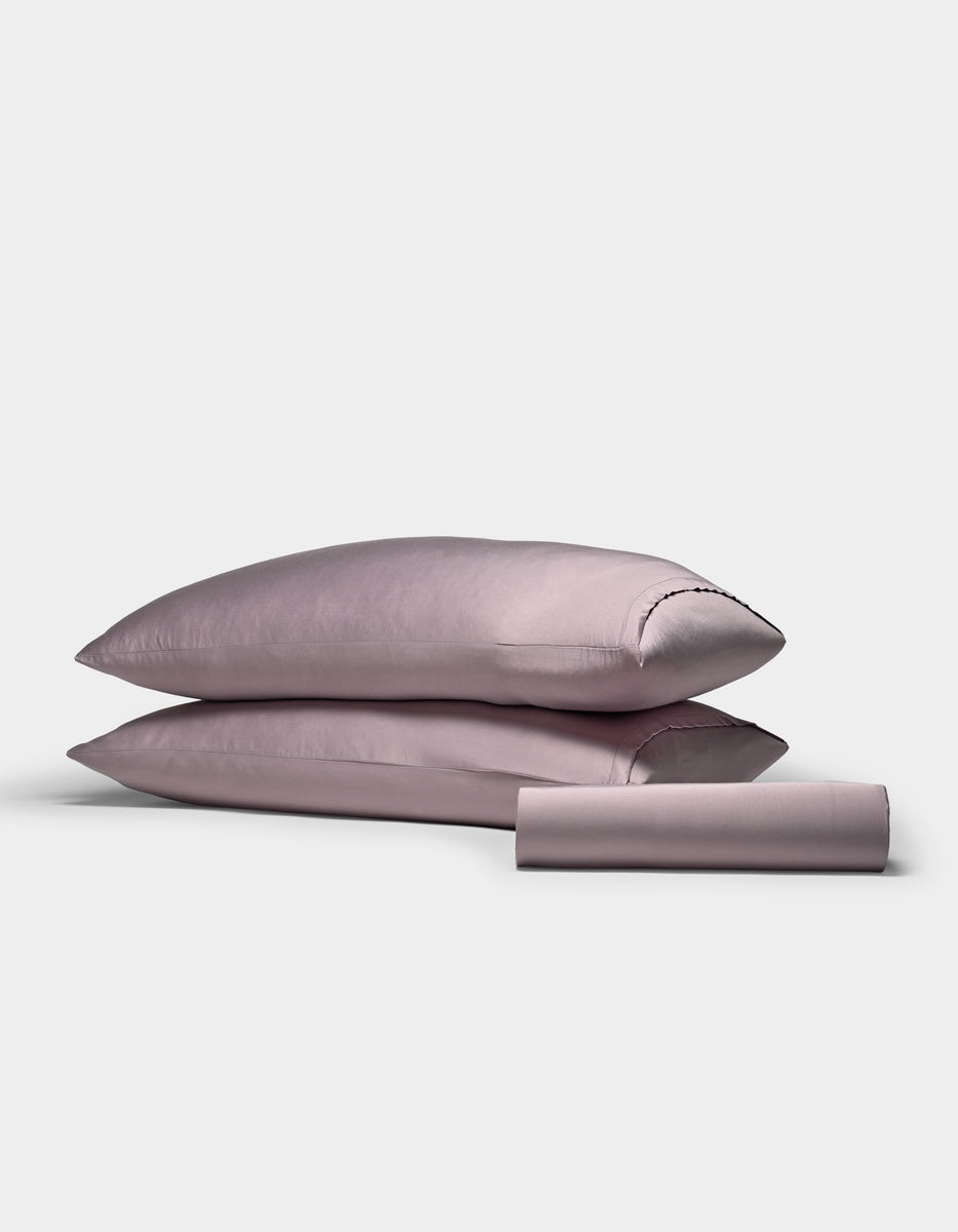 Two stacked lavender pillows and a matching rolled-up sheet from the Cozy Earth Bamboo Bedding Basic Bundle are set against a plain white background.