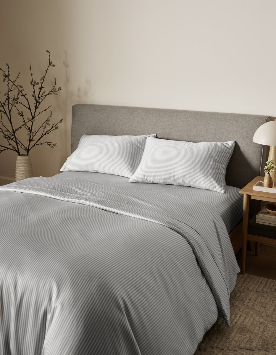A neatly made bed featuring the Cozy Earth Bamboo Duvet Cover in gray and white stripes, styled with two pillows, a gray upholstered headboard, a small bedside table with a lamp, and a vase of branches in a cozy, neutral-toned bedroom.