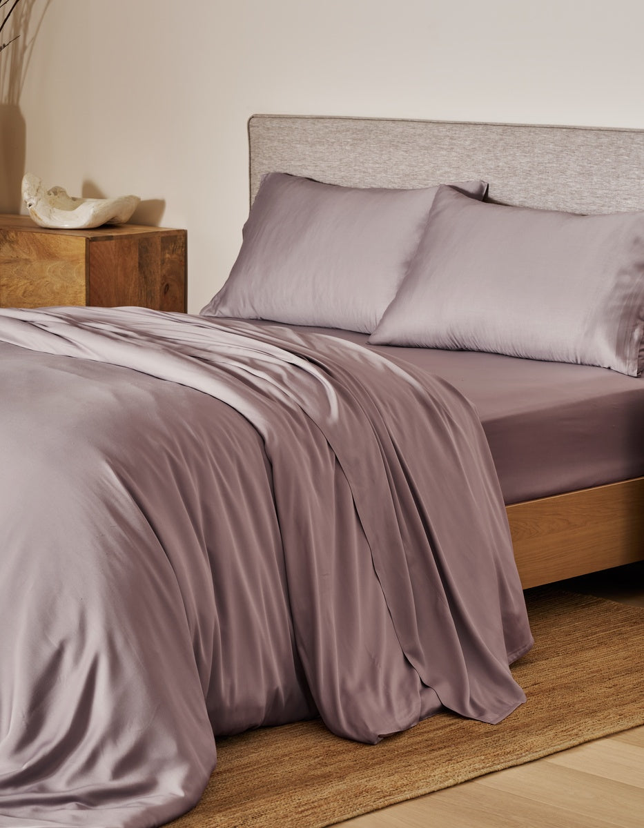 A neatly made bed featuring Cozy Earth’s light mauve Bamboo Duvet Cover and matching pillowcases, complemented by a wooden nightstand adorned with a decorative shell, and a neutral headboard, all set atop a woven rug in a softly lit bedroom.