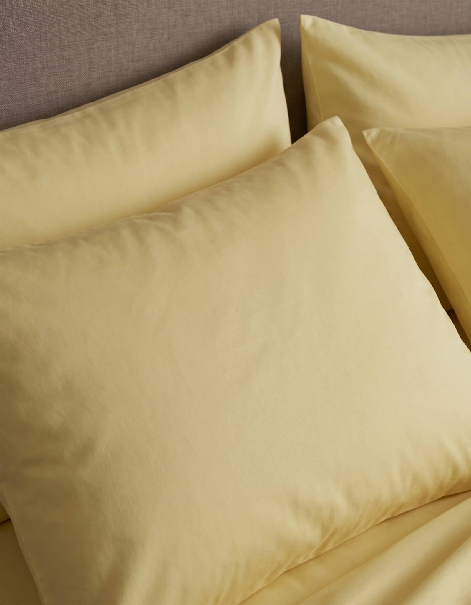 Close-up of neatly arranged Cozy Earth Bamboo Jersey Pillowcases in pale yellow on a bed with a beige upholstered headboard.