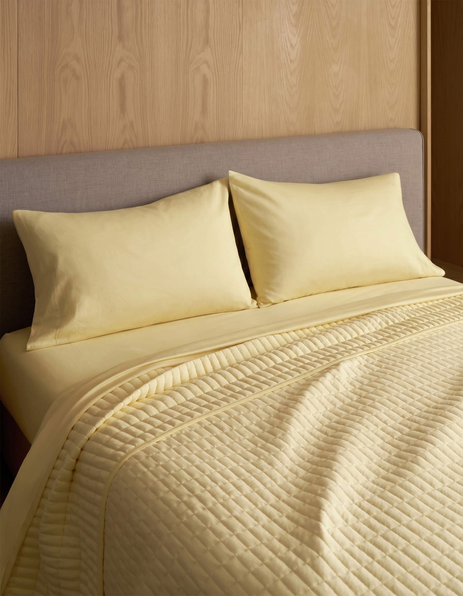 A neatly made bed features Cozy Earth Bamboo Jersey Pillowcases, two matching pillows, and a quilted yellow blanket, all set against a wood-paneled wall and a light gray upholstered headboard.