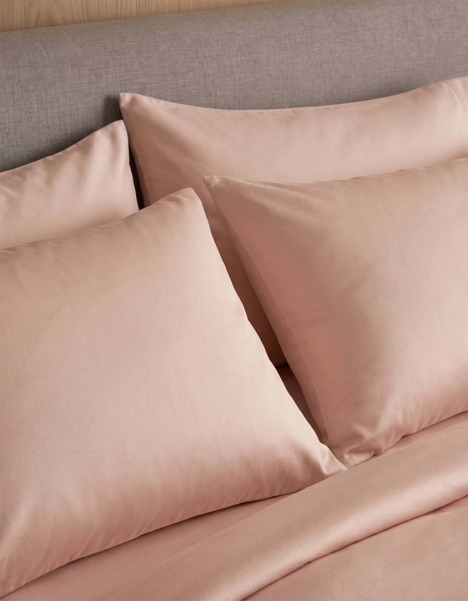 A close-up of a neatly made bed with Cozy Earth Bamboo Jersey Pillowcases in light pink, paired with matching sheets and set against a grey upholstered headboard.