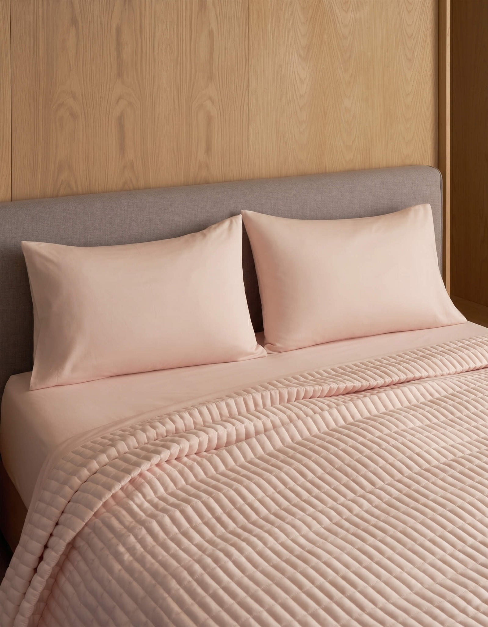A neatly made bed features two Cozy Earth Bamboo Jersey Pillowcases in light pink, matching sheets, a quilted pink blanket, and a gray upholstered headboard set against a wooden wall.