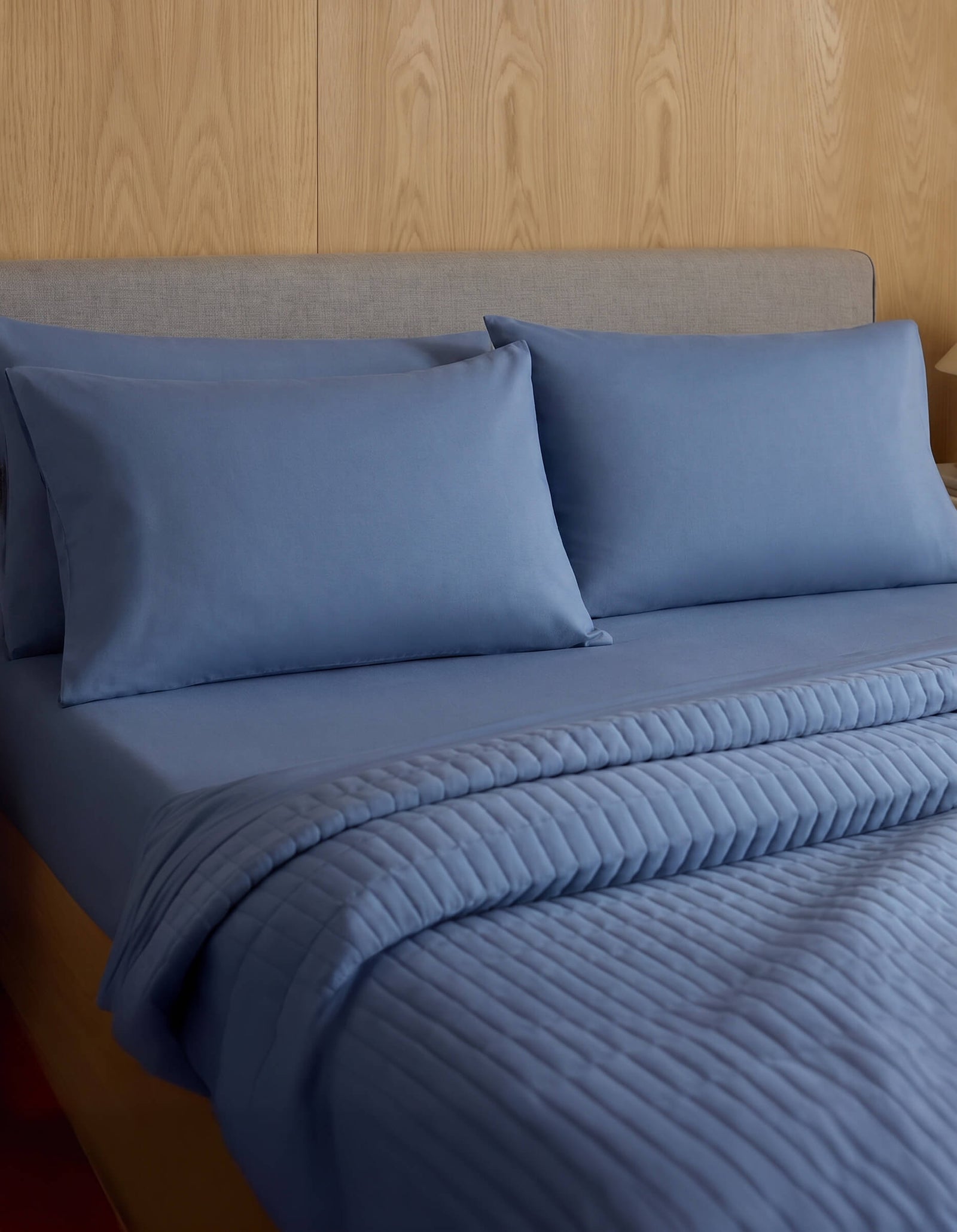 A neatly made bed features Cozy Earth Bamboo Jersey Pillowcases, matching pillows, and a quilted blue blanket, all set against a wood-paneled wall and a light gray upholstered headboard.