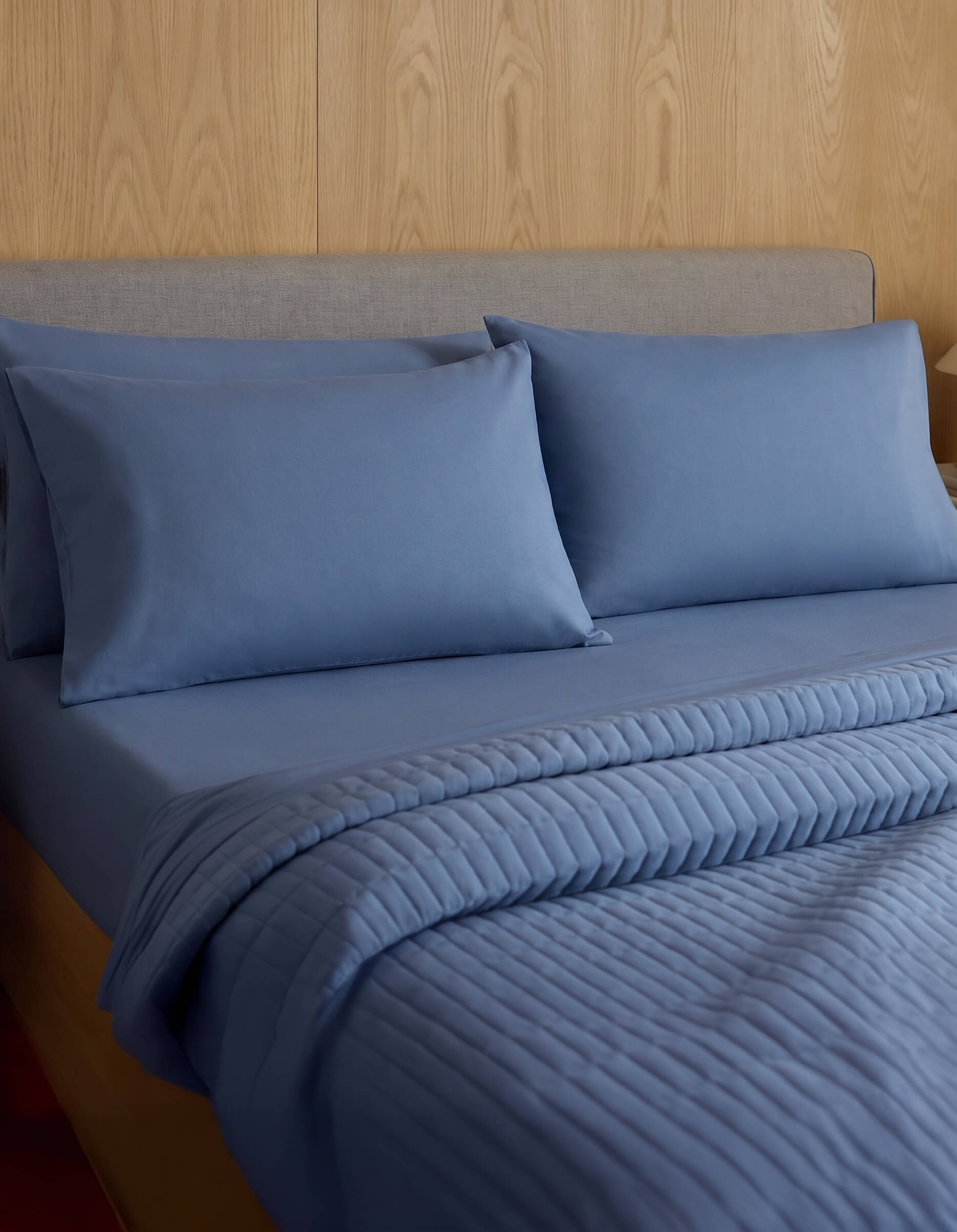 A neatly made bed features Cozy Earth Bamboo Jersey Pillowcases, matching pillows, and a quilted blue blanket, all set against a wood-paneled wall and a light gray upholstered headboard.