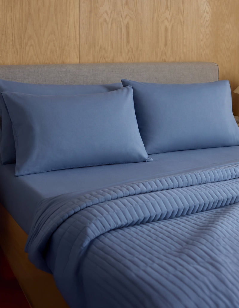 A neatly made bed features Cozy Earth Bamboo Jersey Pillowcases, matching pillows, and a quilted blue blanket, all set against a wood-paneled wall and a light gray upholstered headboard.