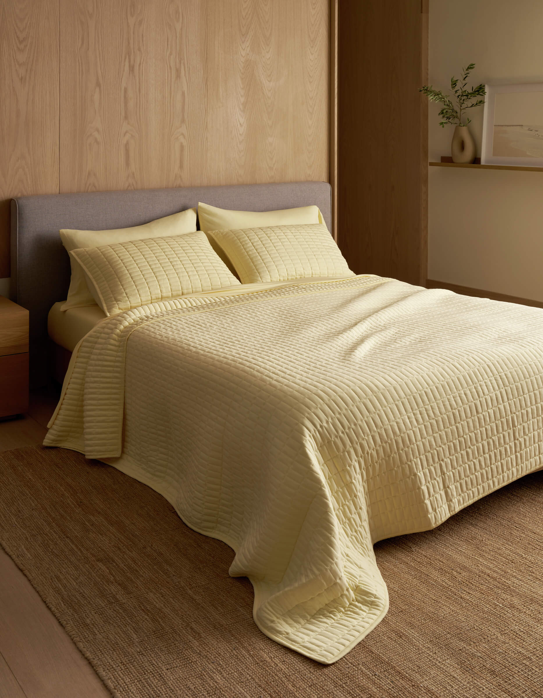 A minimalist bedroom features a neatly made bed with the Cozy Earth Bamboo Jersey Quilt in pale yellow, matching pillows, wood-paneled walls, a neutral rug, and a small shelf displaying a vase and framed art.