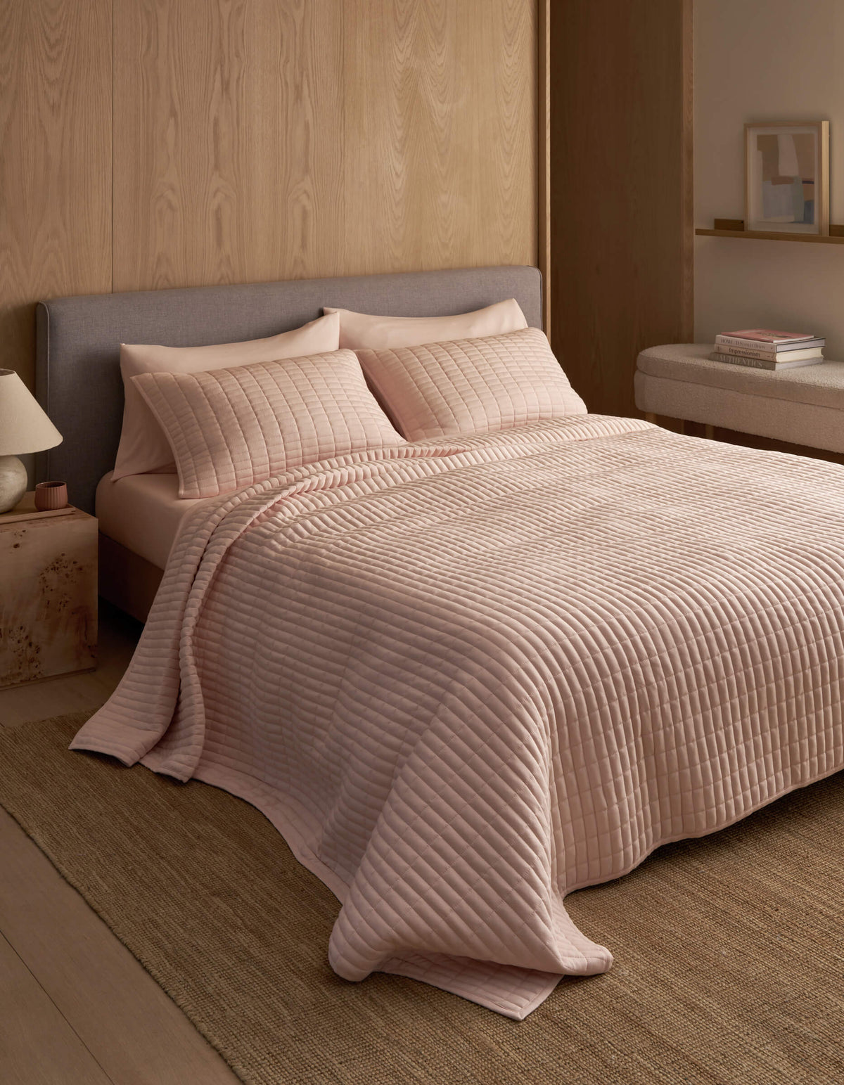 A neatly made bed features the Cozy Earth Bamboo Jersey Quilt in light pink with matching pillows, set against a wooden wall in a minimal, modern bedroom with neutral tones and simple decor.|Color:Petal