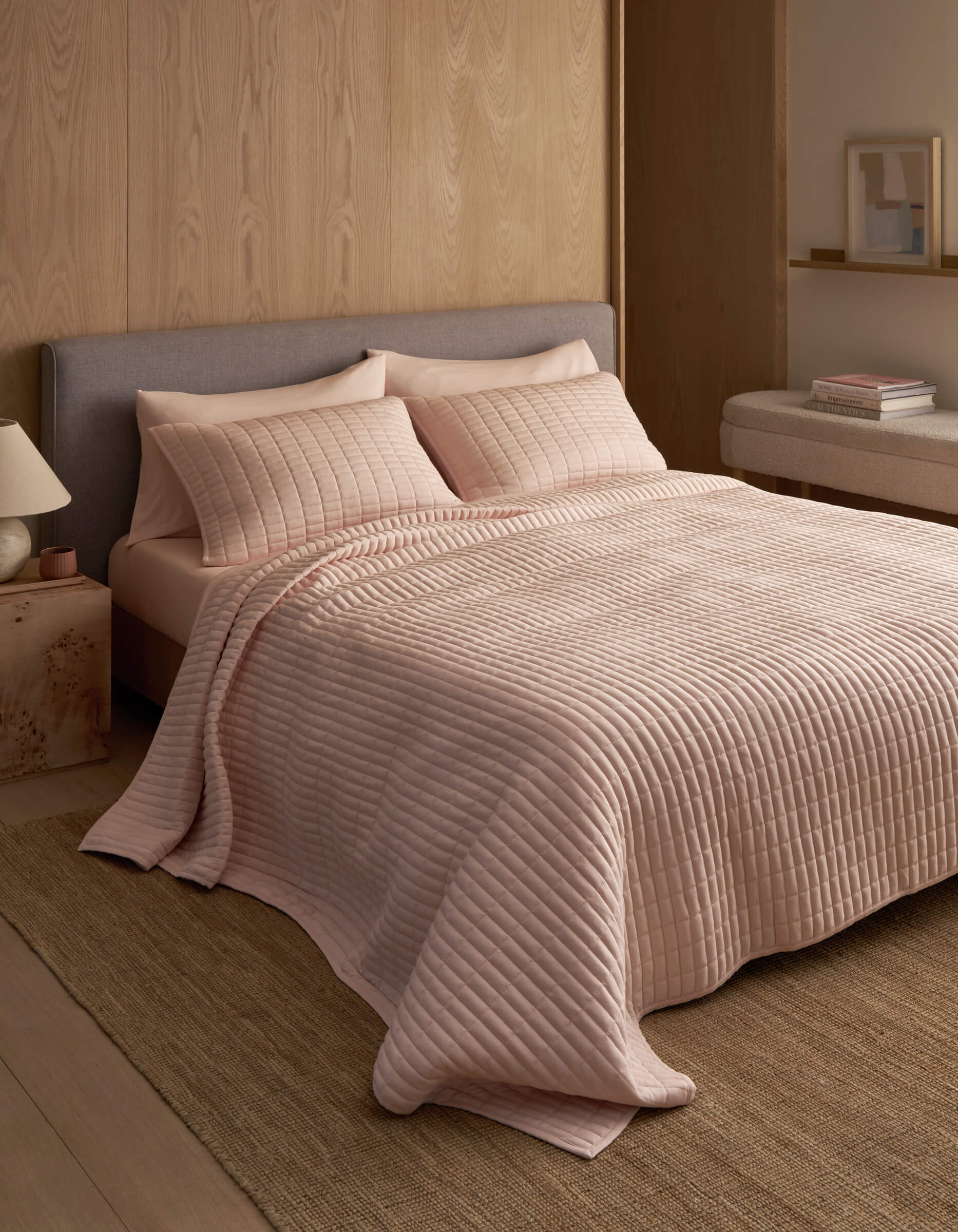 A neatly made bed features the Cozy Earth Bamboo Jersey Quilt in light pink with matching pillows, set against a wooden wall in a minimal, modern bedroom with neutral tones and simple decor.|Color:Petal