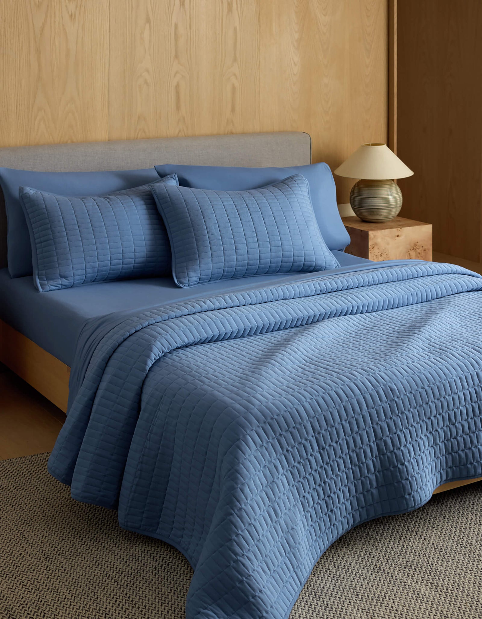 A neatly made bed features the Cozy Earth Bamboo Jersey Quilt with matching pillows, set against a light wood-paneled wall. A small nightstand with a lamp sits beside the bed on a textured rug.