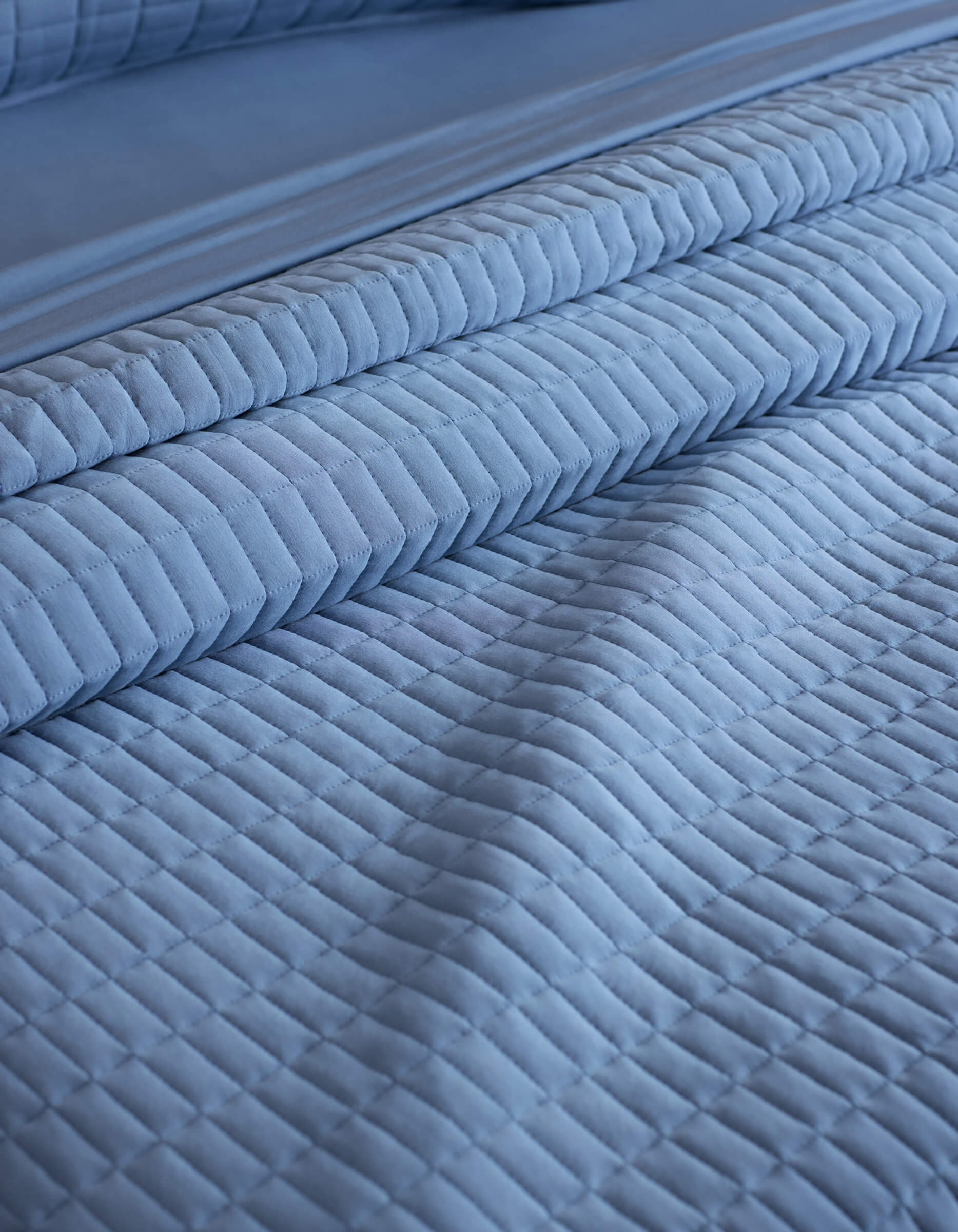 Close-up of a neatly made bed with the Cozy Earth Bamboo Jersey Quilt in light blue, featuring a quilted grid pattern and matching pillowcases. The soft, textured fabric creates an inviting look.|Color:Sailor