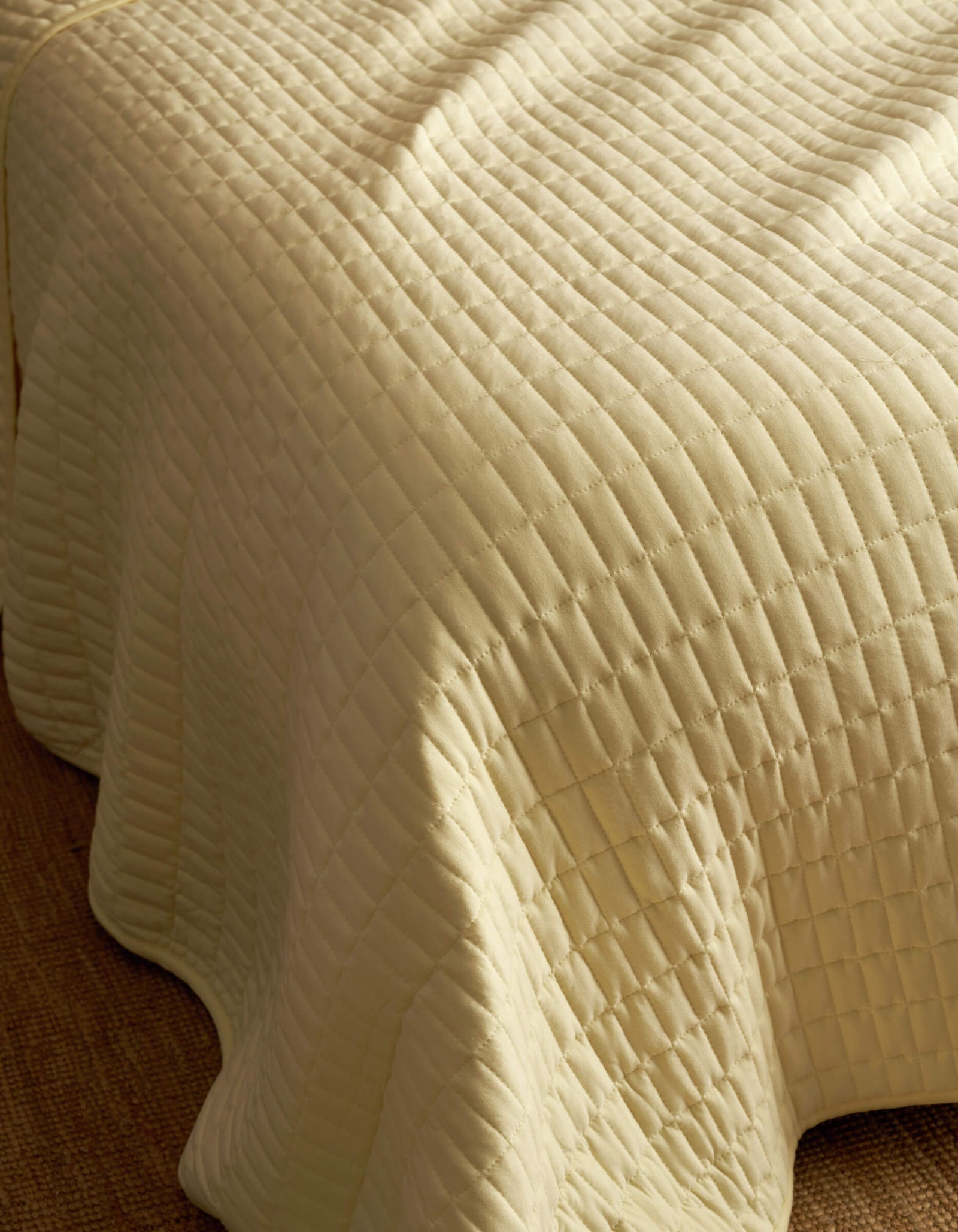 A close-up of the Bamboo Jersey Quilt by Cozy Earth, featuring a cream color, quilted square pattern, and soft textured stitching draped over the edge of a bed.