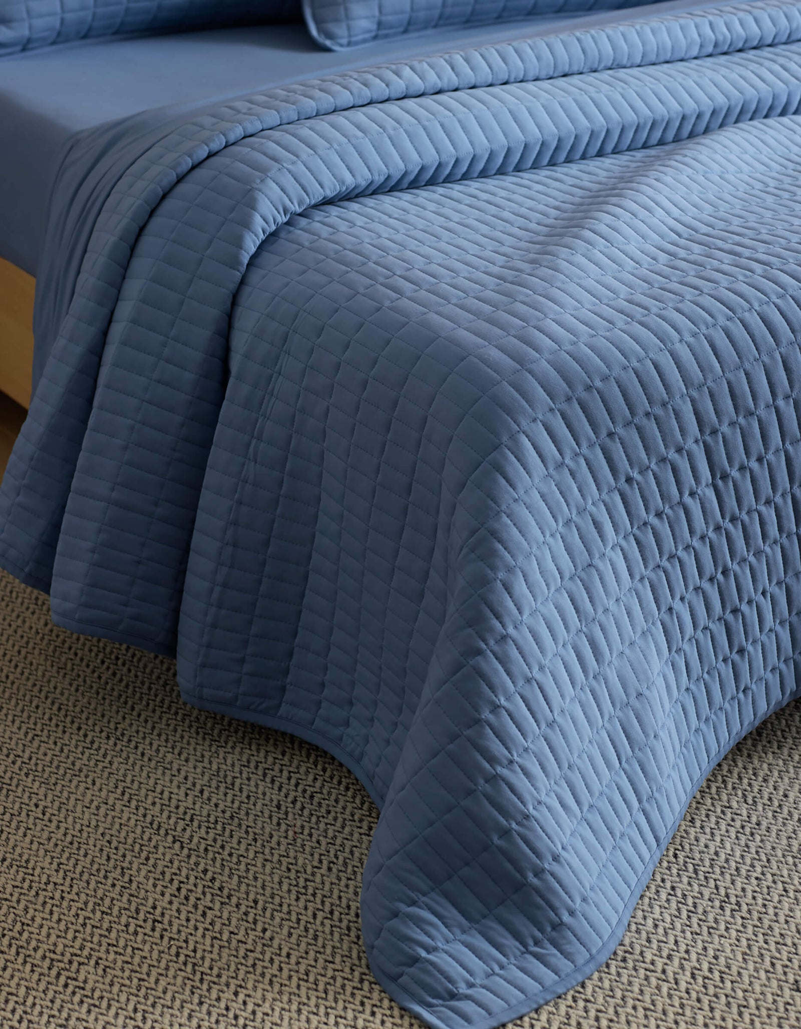 A close-up of a neatly made bed features the Cozy Earth Bamboo Jersey Quilt, its textured blue surface draped elegantly over the edge and resting on a beige woven carpet.