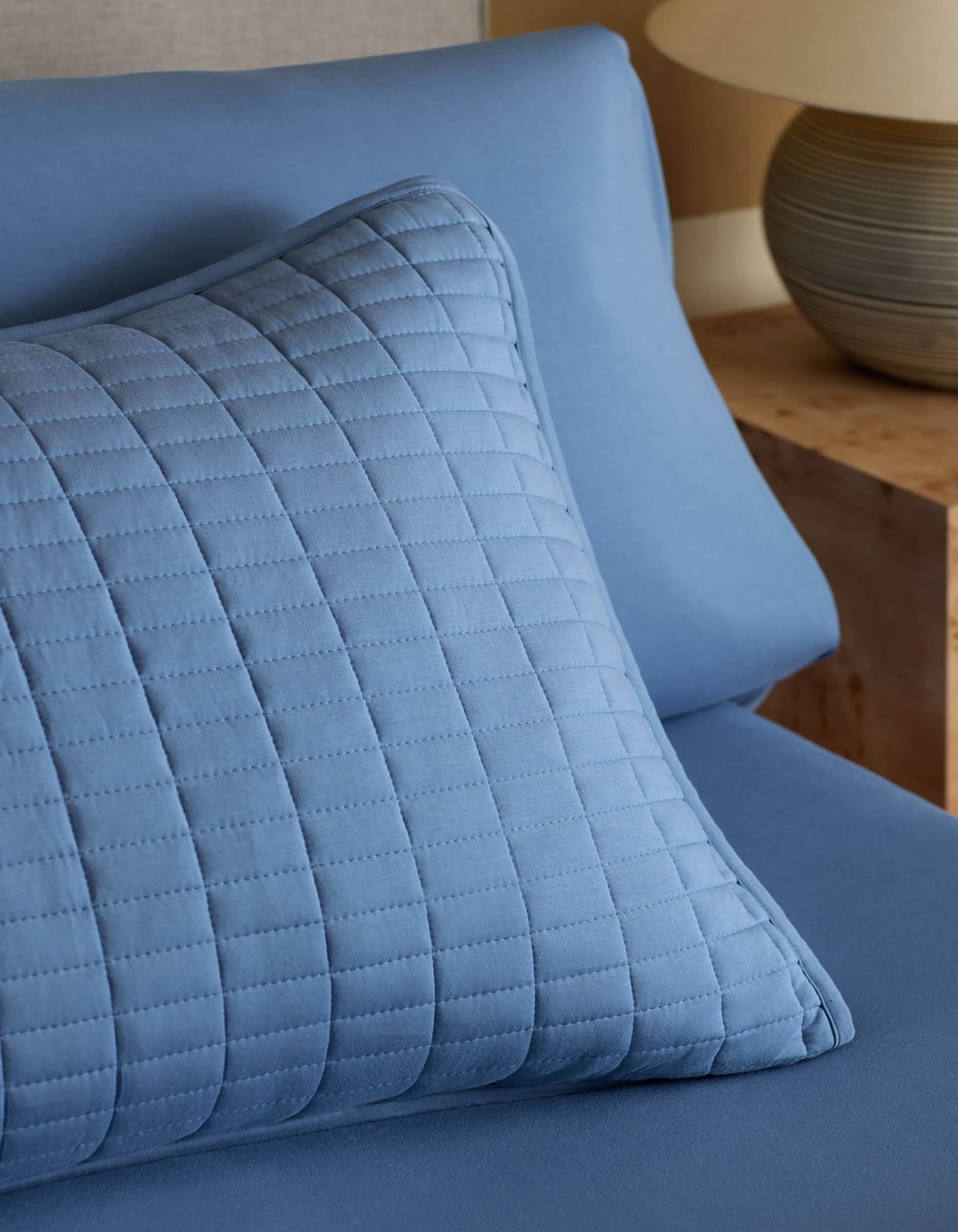 A Cozy Earth Bamboo Jersey Quilted Sham in blue rests on matching sheets, with a wooden nightstand and ceramic lamp visible in the background.
