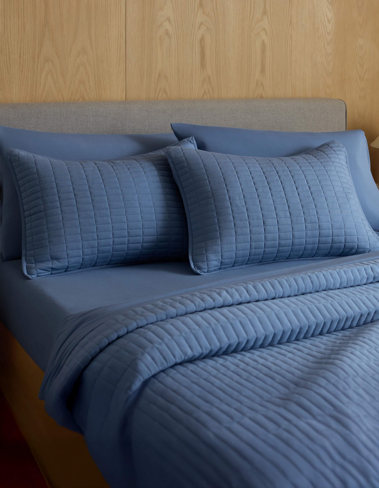 A neatly made bed with a grey headboard features Cozy Earth's Bamboo Jersey Quilted Shams in blue, a matching fitted sheet, and comforter, all set against a light wood-paneled wall.