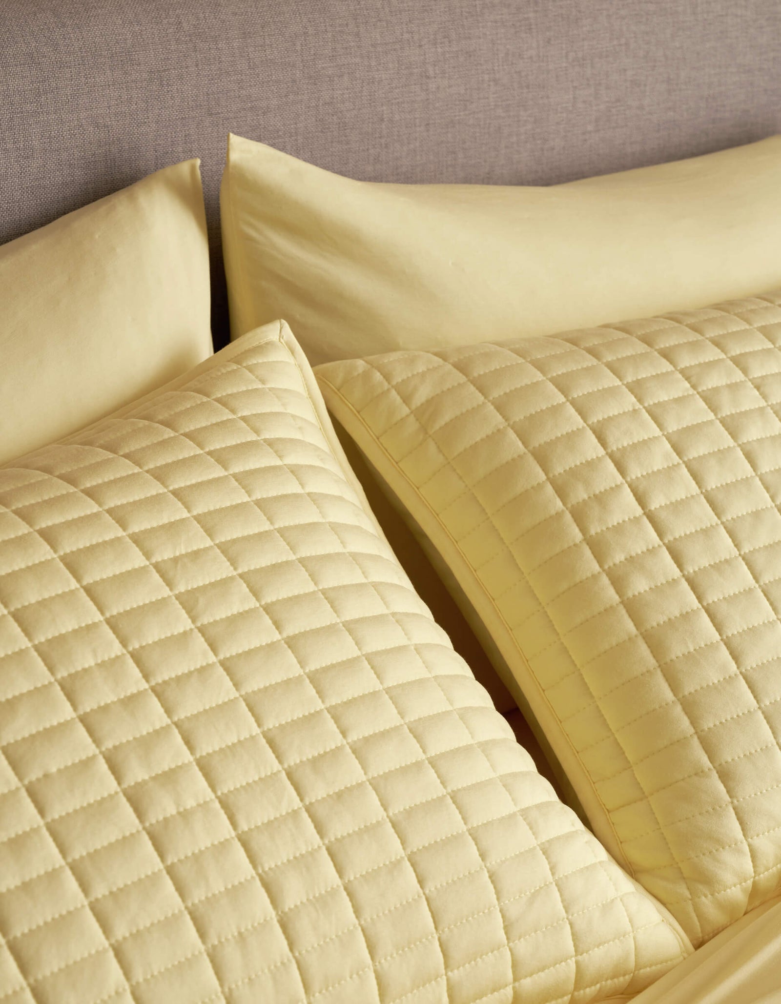 Two Cozy Earth Bamboo Jersey Quilted Shams in yellow, featuring a square pattern, rest against a grey upholstered headboard, paired with matching yellow bedsheets and pillowcases.