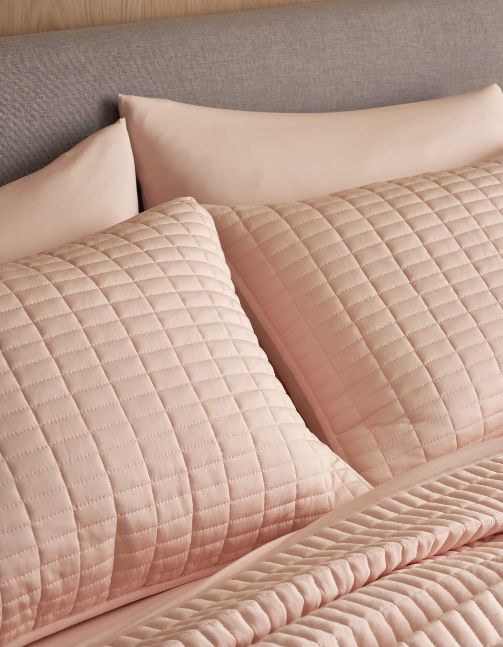 Close-up of a neatly made bed featuring Cozy Earth's Bamboo Jersey Quilted Shams and matching pillowcases in light pink, paired with a quilted bedspread. The upholstered headboard is covered in soft gray fabric.