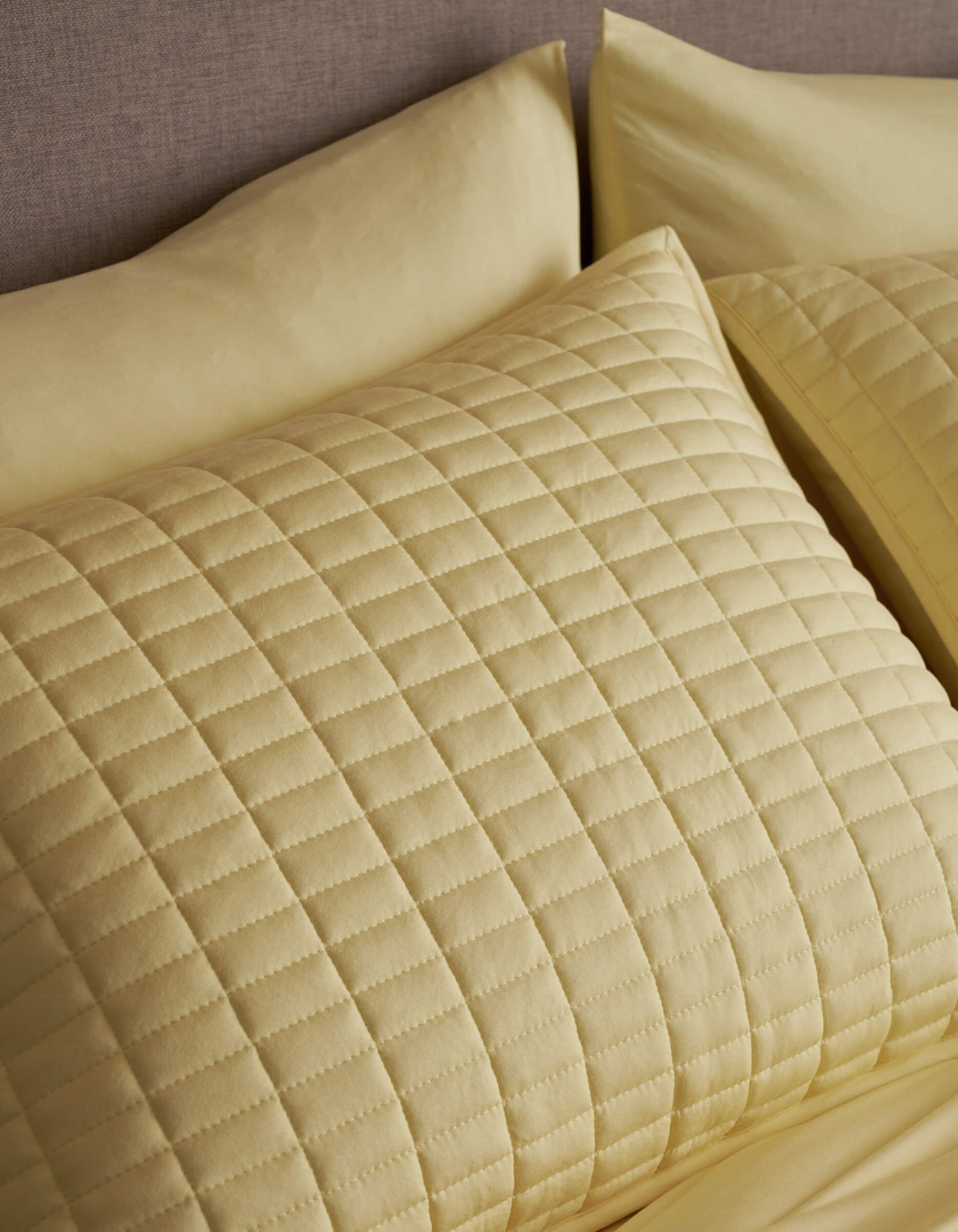 A close-up of a neatly made bed showcases Cozy Earth's Bamboo Jersey Quilted Shams in light yellow, featuring a square pattern and soft, textured appearance that matches the sheets.
