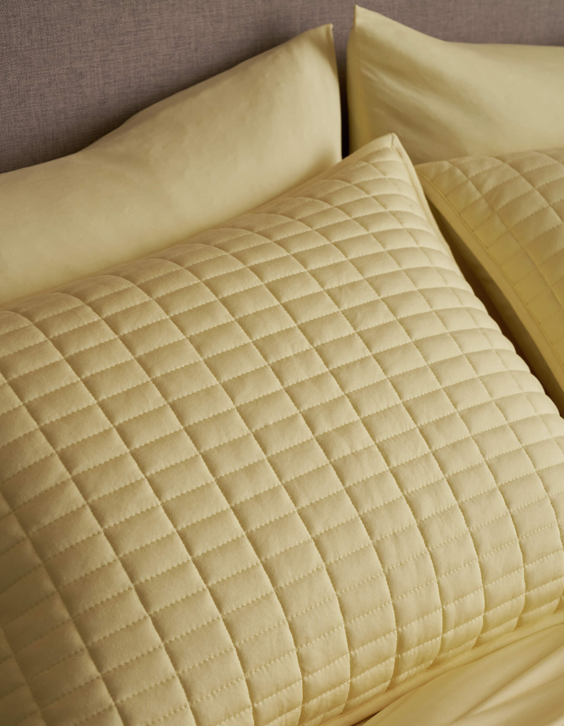 A close-up of a neatly made bed showcases Cozy Earth's Bamboo Jersey Quilted Shams in light yellow, featuring a square pattern and soft, textured appearance that matches the sheets.