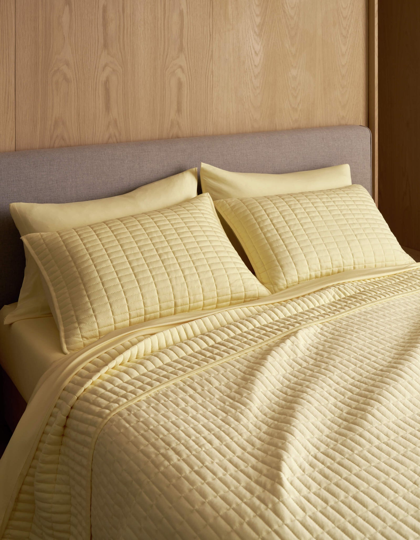 A neatly made bed features the Cozy Earth Bamboo Jersey Quilt in light yellow with matching pillow shams, set against a light wood paneled wall and a gray upholstered headboard.