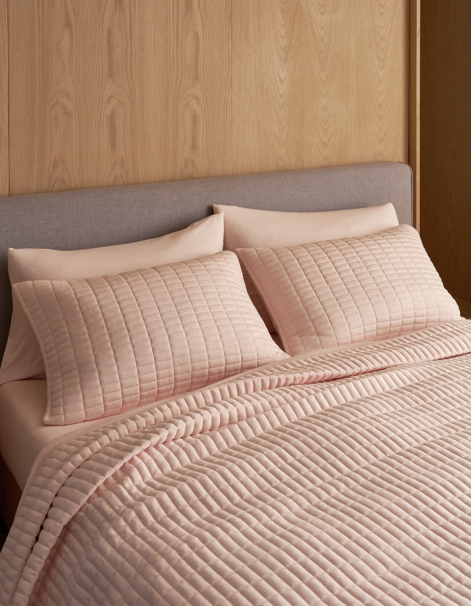 A neatly made bed features the Cozy Earth Bamboo Jersey Quilt in light pink with matching pillows, set against a light wood-paneled wall and a light grey upholstered headboard.