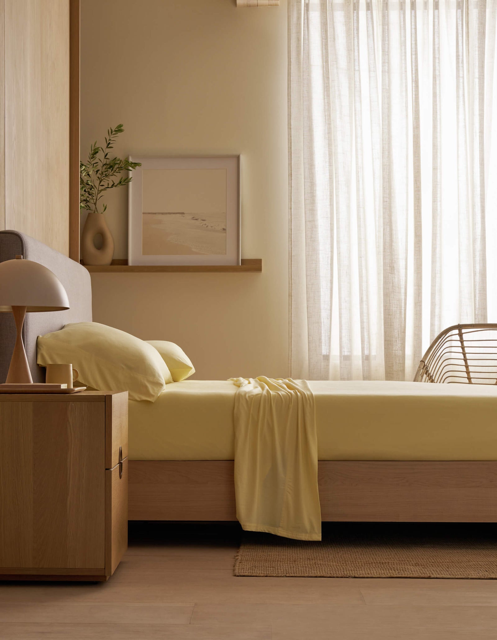 A minimal bedroom with a wooden bed frame, Cozy Earth's Bamboo Jersey Sheet Set in pale yellow, a wooden nightstand and lamp, a plant, framed art on a shelf, and sheer curtains letting in natural light.