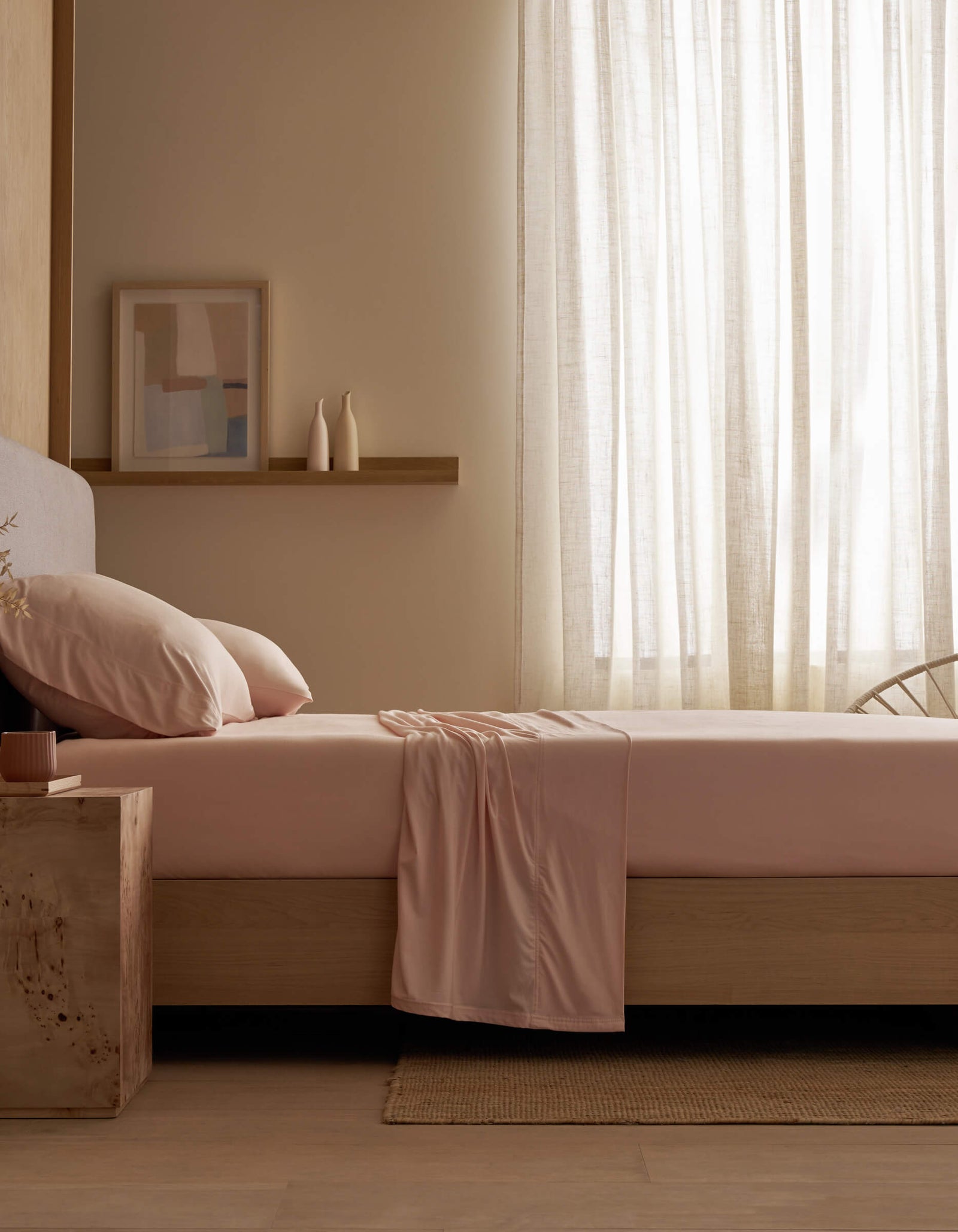 A minimal bedroom features a neatly made bed with Cozy Earth's Bamboo Jersey Sheet Set in pale pink, soft light filtered through sheer curtains, a wooden side table, framed abstract art, and two vases on a wall shelf.