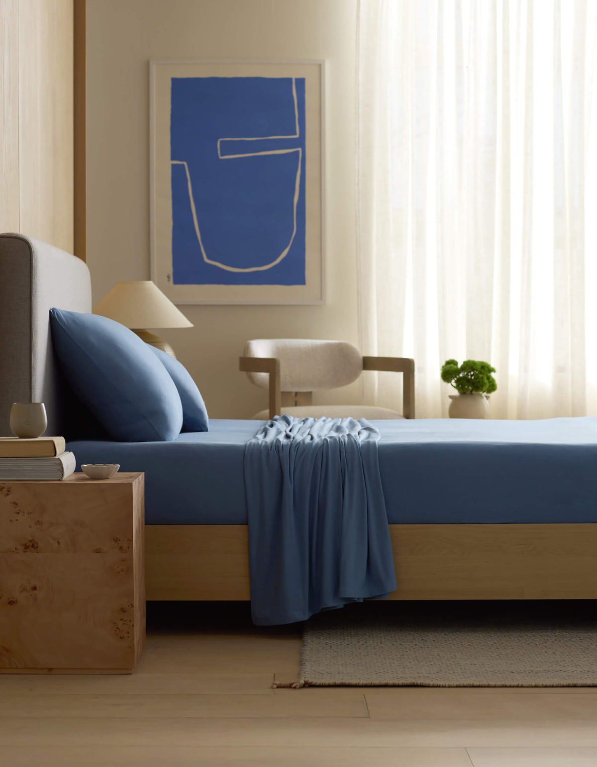 A modern bedroom features a bed with Cozy Earth’s Bamboo Jersey Sheet Set in blue, a light wood nightstand with a cup, books, and lamp, a chair, a plant, sheer curtains, abstract blue wall art, and soft natural light.|Color:Sailor