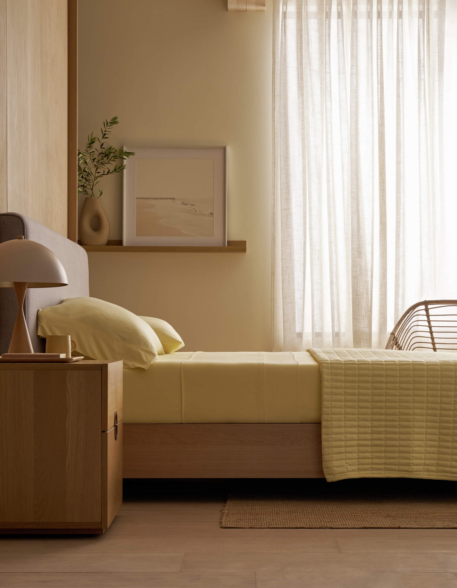 A minimalist bedroom with light wood furniture, Cozy Earth Bamboo Jersey Pillowcases on the bed, yellow bedding, a nightstand with a lamp, a plant, wall art, and soft light through sheer white curtains.