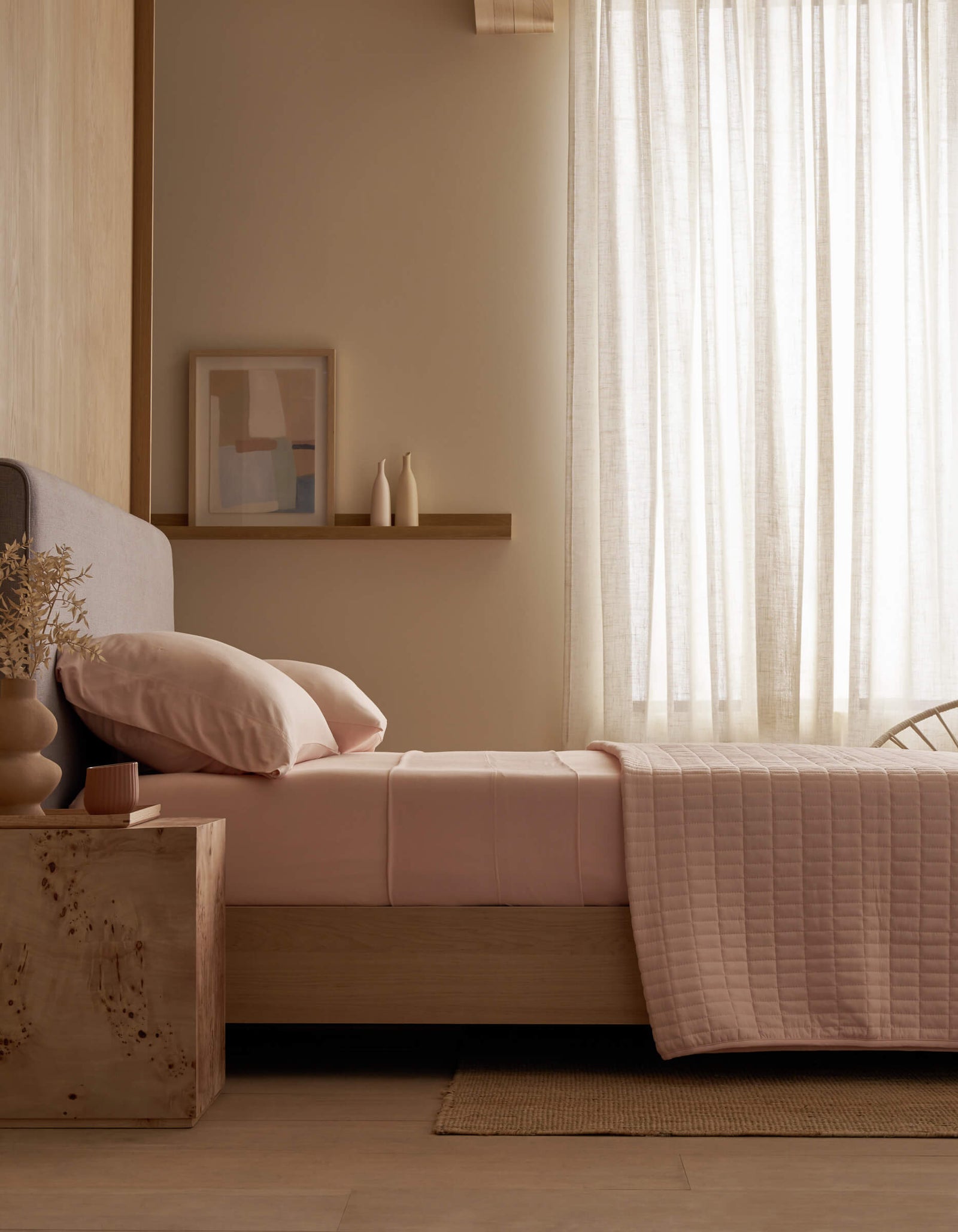 A modern bedroom featuring a bed with Cozy Earth Bamboo Jersey Fitted Sheet in pink, a wooden nightstand with books and a cup, a beige chair, potted plant, sheer curtains, and abstract blue wall art.
