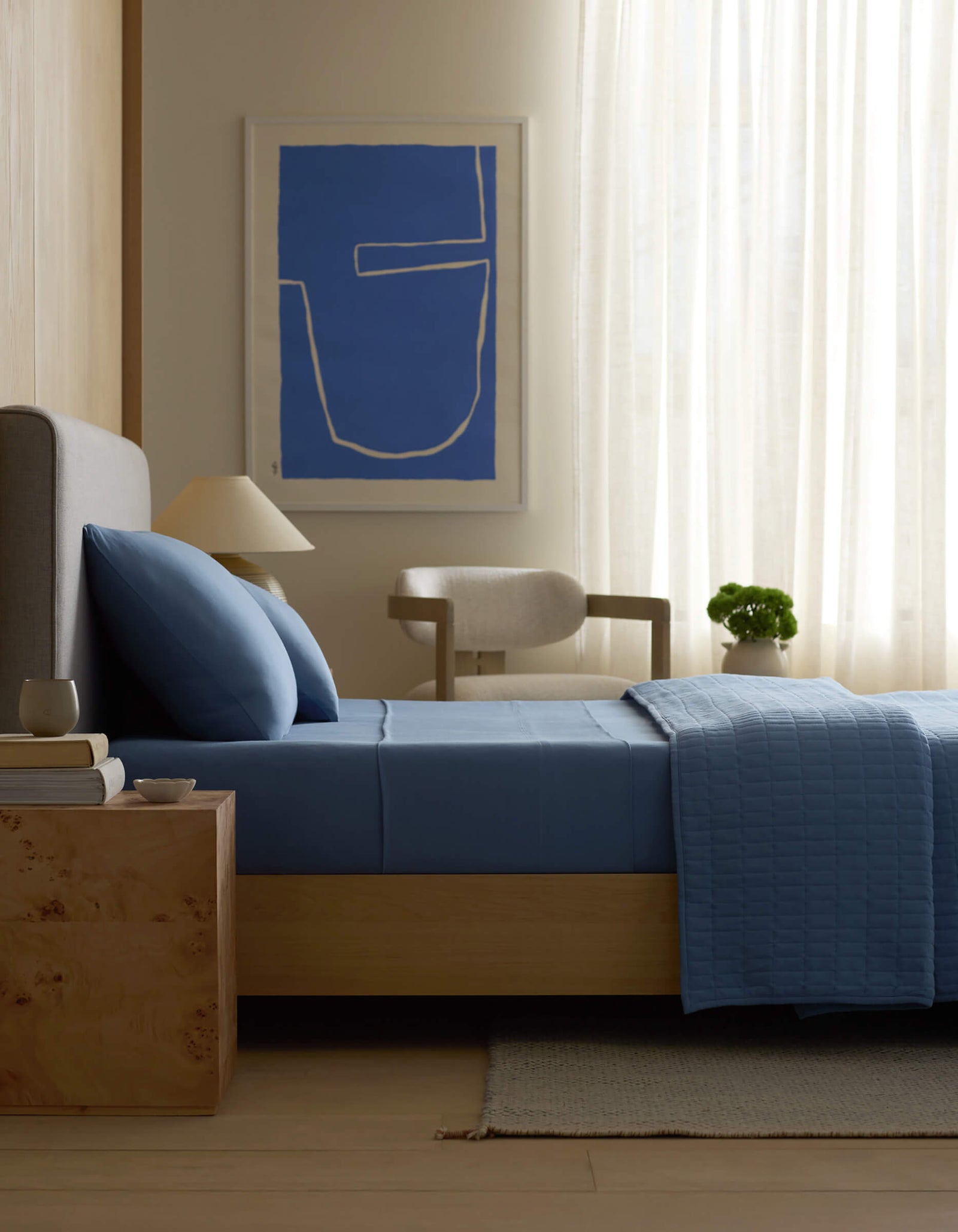 A modern bedroom featuring a bed with Cozy Earth Bamboo Jersey Fitted Sheet in blue, a wooden nightstand with books and a cup, a beige chair, potted plant, sheer curtains, and abstract blue wall art.