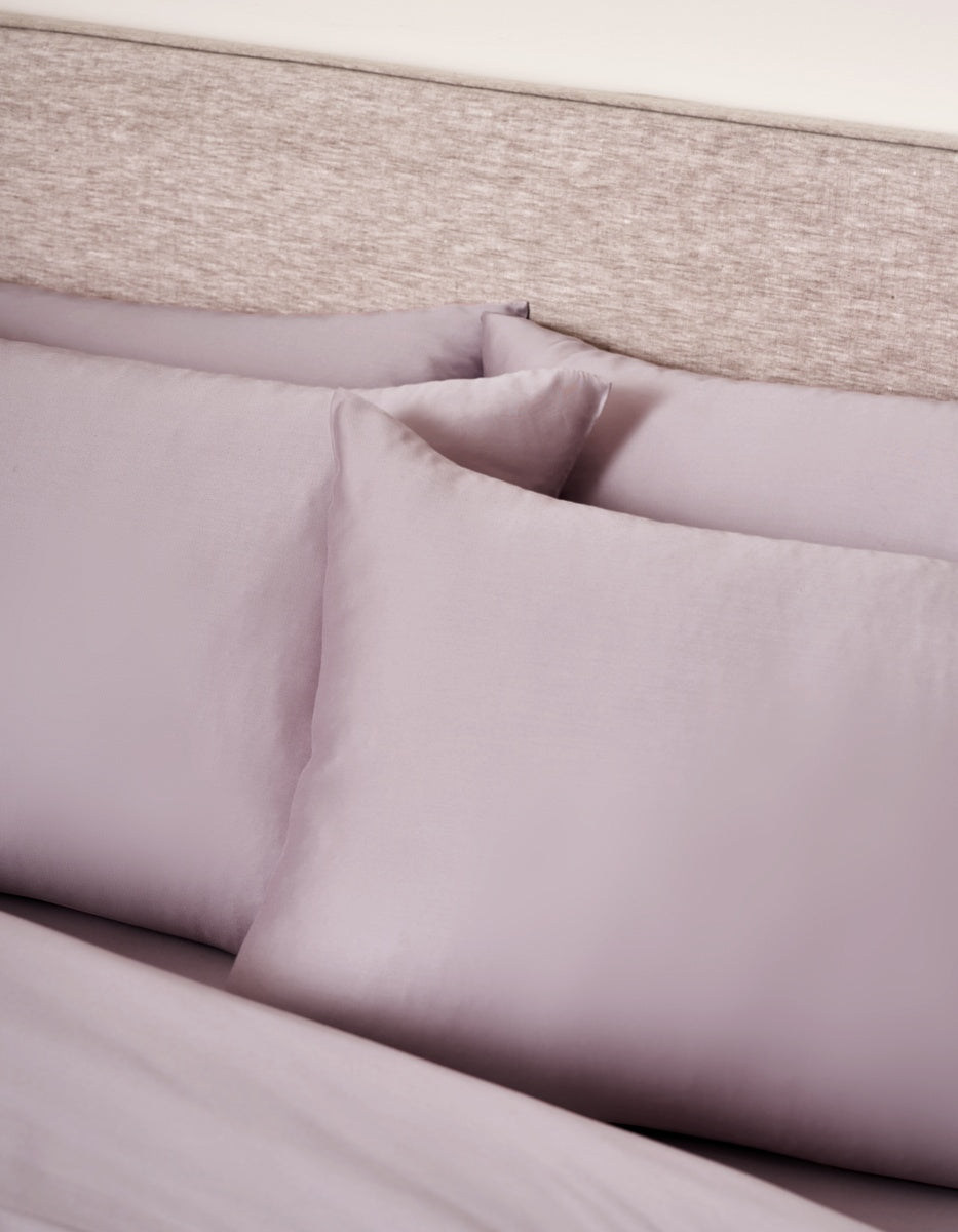 Two Cozy Earth Bamboo Pillowcases in lavender rest against a matching headboard, with a coordinating lavender sheet partially visible beneath them. The bedding and pillowcases feature a smooth, soft texture.