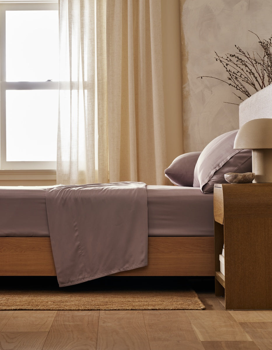 A neatly made bed is dressed in Cozy Earth’s Bamboo Flat Sheet, paired with soft pillows and positioned next to a wooden nightstand topped with a lamp and decorative branches. Light filters through sheer curtains, creating a calm, cozy bedroom atmosphere.