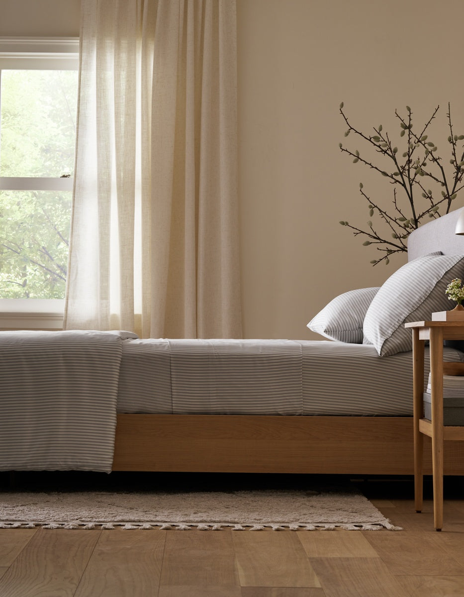 A neatly made bed featuring the Cozy Earth Bamboo Fitted Sheet sits beside a wooden nightstand, a branch in a vase, and a window with sheer curtains letting in natural light. Light wood floors and a rug complete the cozy scene.