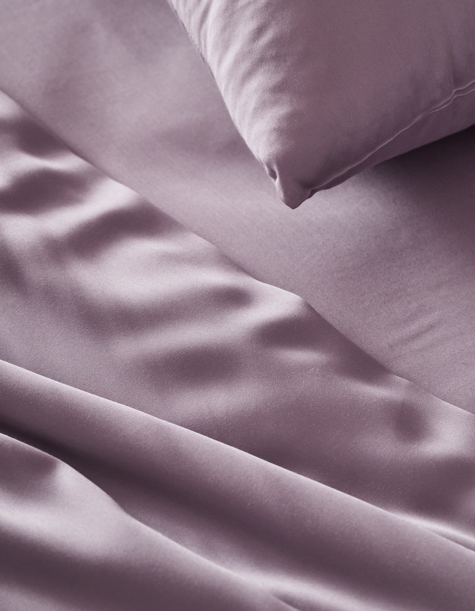 A close-up of a smooth, shiny Bamboo Flat Sheet and matching pillowcase by Cozy Earth in lavender, showcasing soft folds and a luxurious texture.
