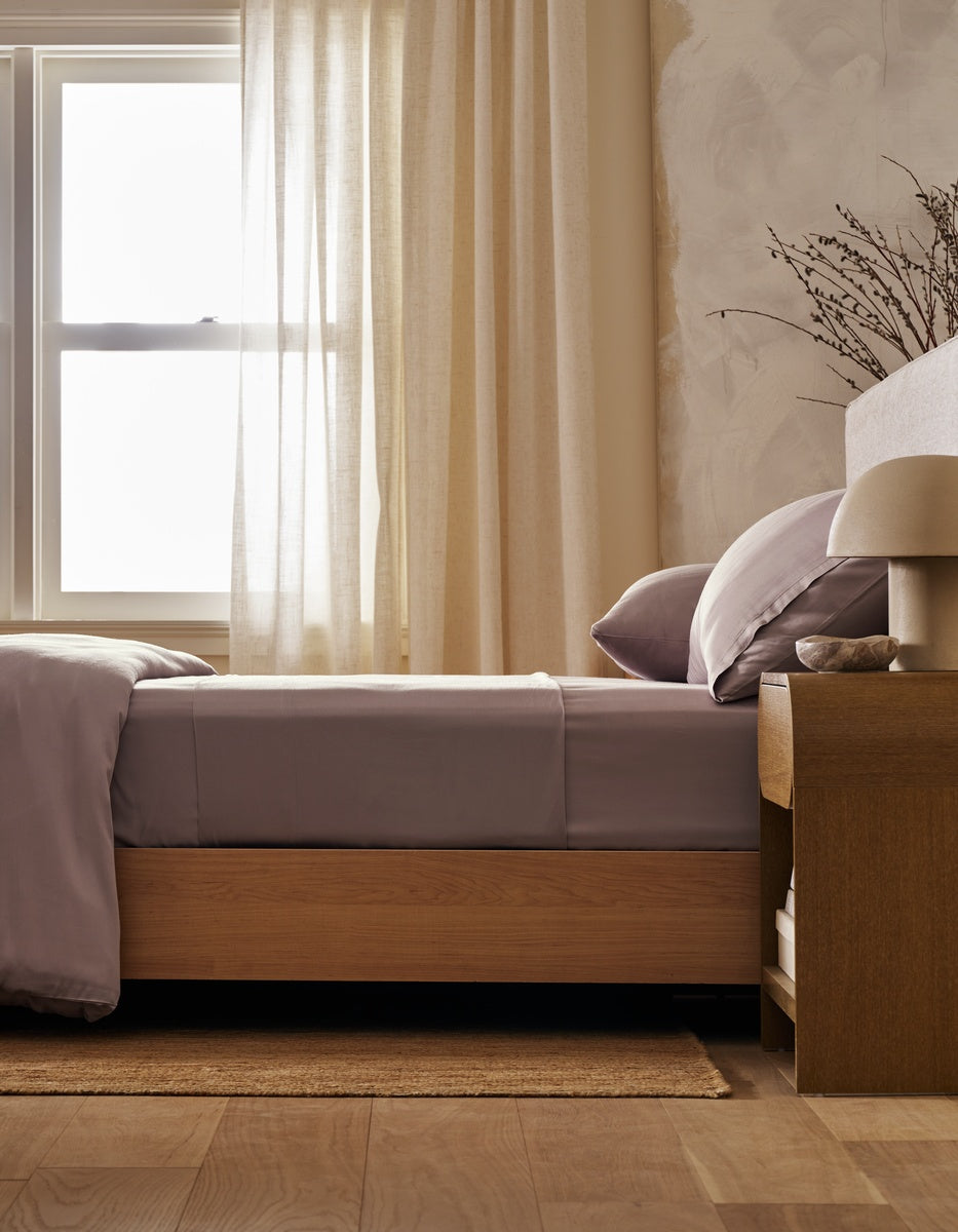 A neatly made bed with Cozy Earth’s Bamboo Fitted Sheet sits beside a wooden nightstand adorned with a lamp and dried branches, while light curtains allow sunlight to filter in through the window.