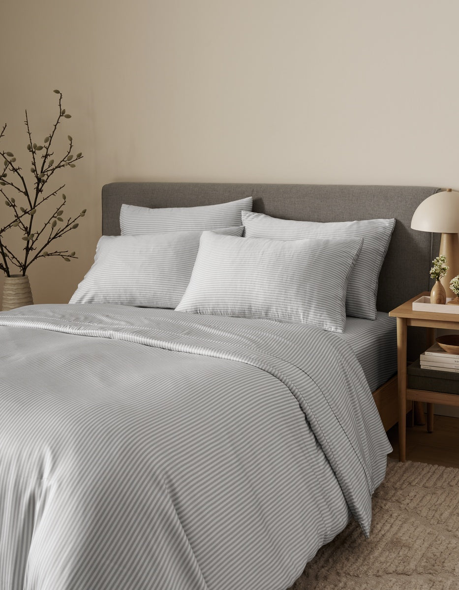 A neatly made bed features the Bamboo Bedding Ultimate Bundle by Cozy Earth, showcasing light gray striped linens and a gray upholstered headboard topped with two pillows. Beside the bed, a wooden nightstand holds a lamp, books, and a vase, while a decorative branch accents the corner against a beige wall.|Color:Classic Stripe in Dew