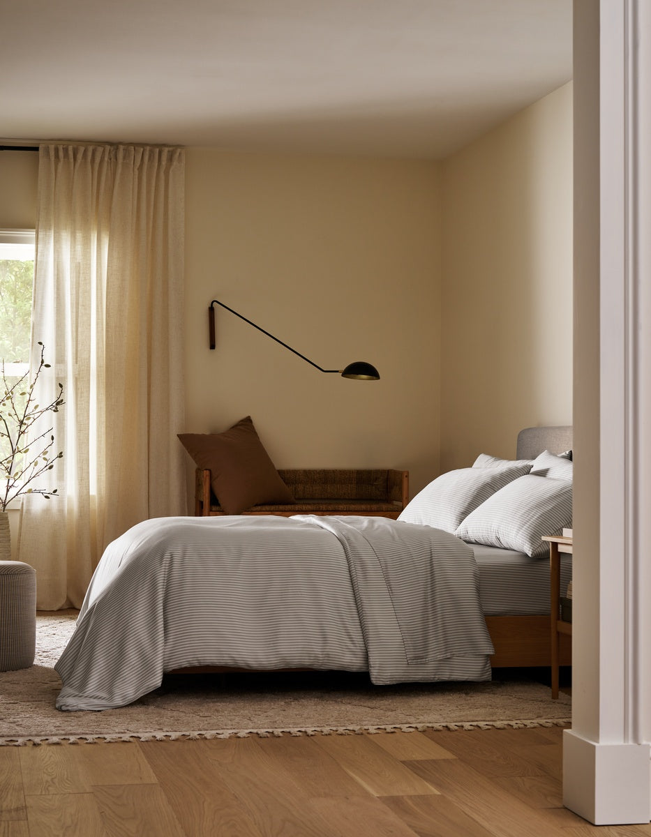 A cozy bedroom featuring a neatly made bed dressed in the Bamboo Bedding Ultimate Bundle by Cozy Earth, complemented by a brown pillow, a wall-mounted lamp, a wooden bench, beige curtains, and light wooden floors. Natural light filters through the window.|Color:Classic Stripe in Dew