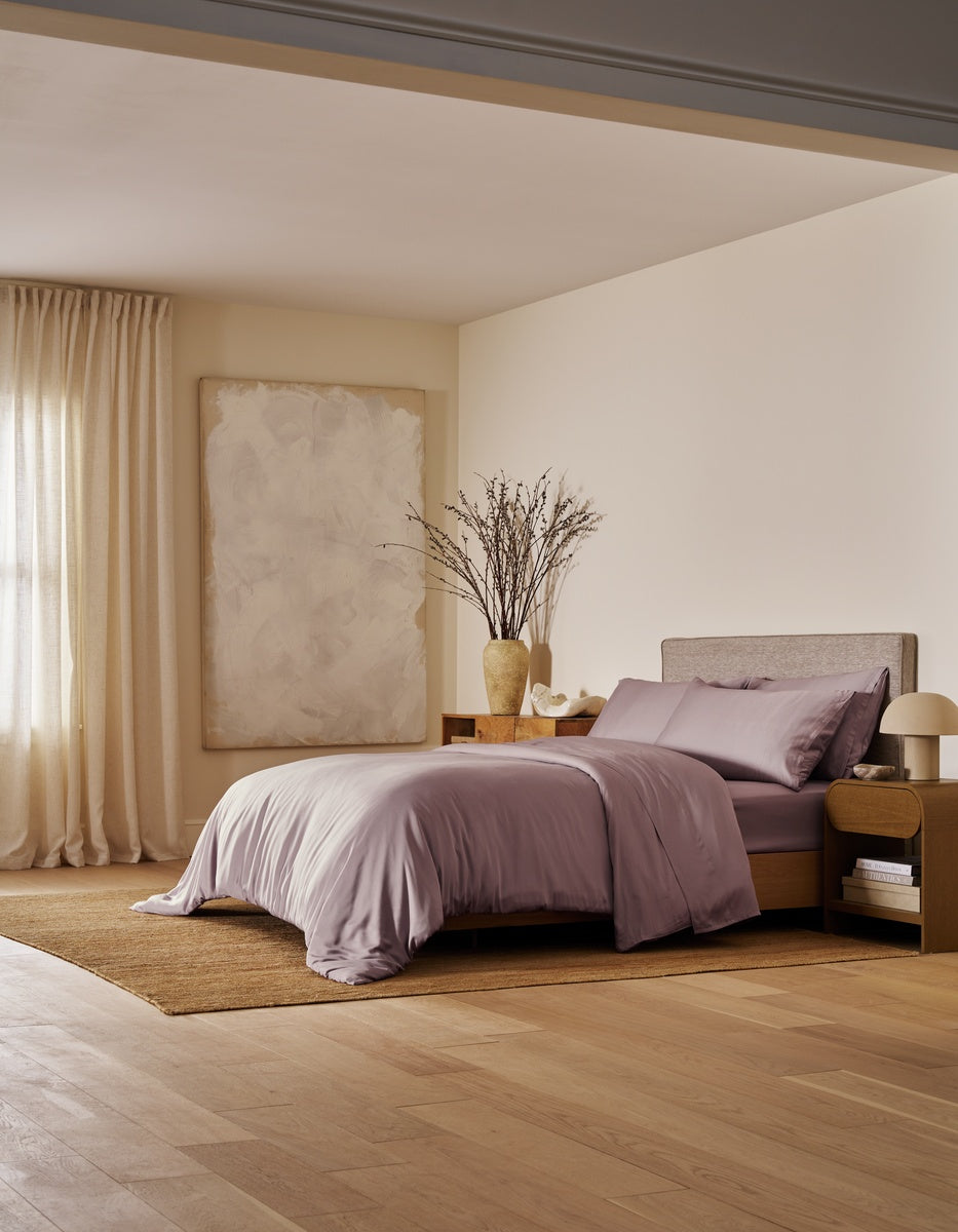 A modern bedroom featuring the Cozy Earth Bamboo Bedding Ultimate Bundle, complemented by neutral walls, large abstract artwork, a floor vase with branches, beige curtains, a wooden nightstand, and bathed in soft natural light.|Color:Lavender