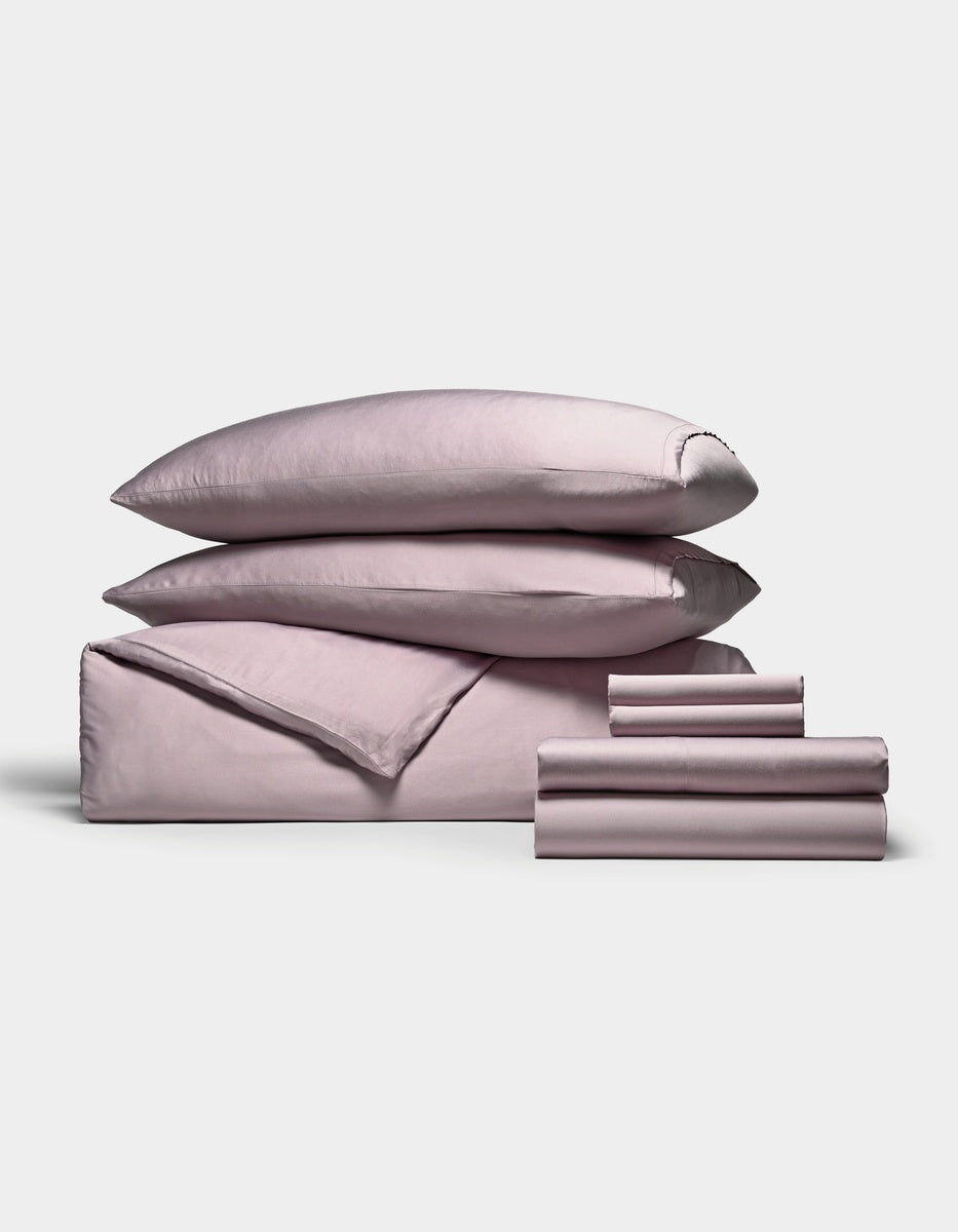 A neatly stacked set of Cozy Earth’s Bamboo Bedding Ultimate Bundle, featuring two pillows, a folded duvet, a flat sheet, and several smaller folded sheets in pale pink, all arranged against a plain white background.