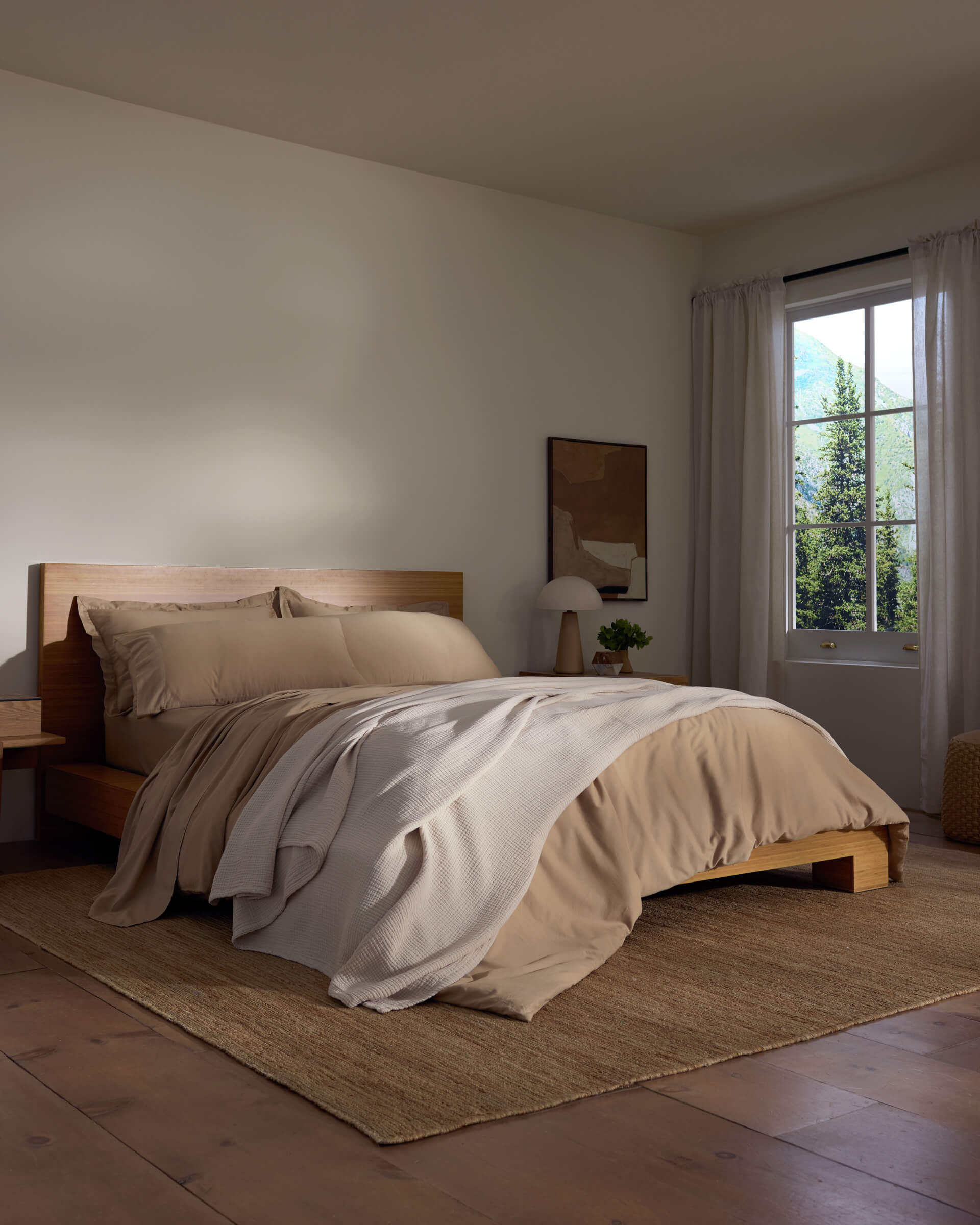 A cozy bedroom features a wooden bed with beige bedding and a white Cotton Gauze Blanket by Cozy Earth, a bedside table with a lamp, sheer curtains letting in natural light, and a woven rug on the wooden floor.