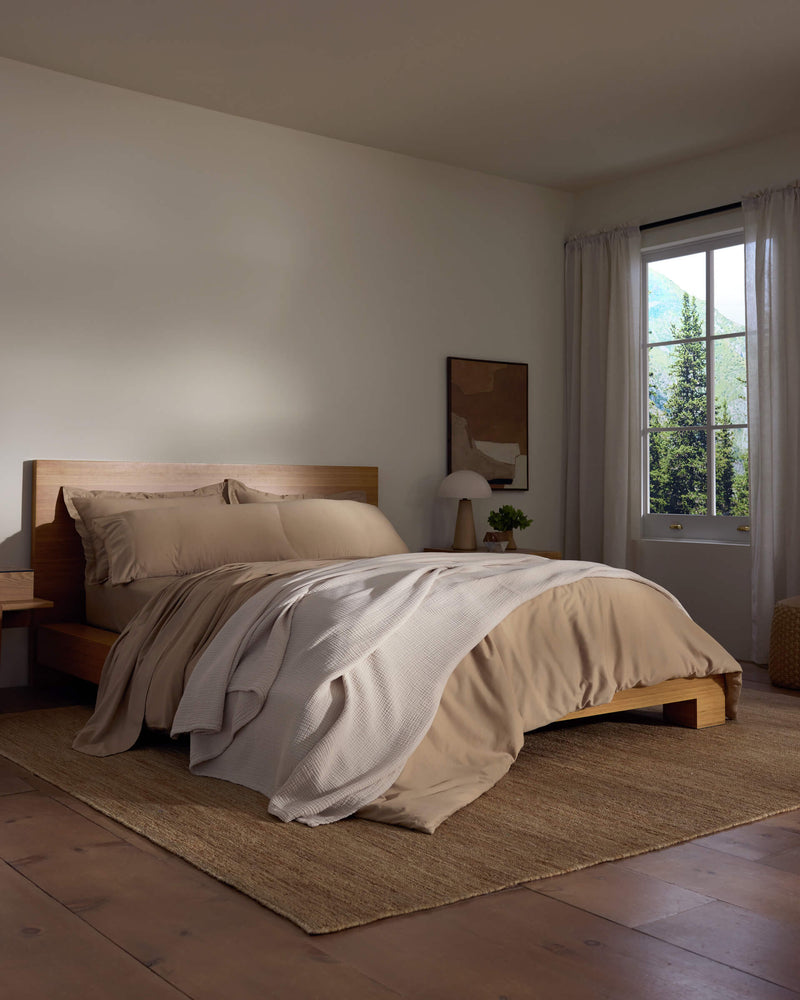 A cozy bedroom features a wooden bed with beige bedding and a white Cotton Gauze Blanket by Cozy Earth, a bedside table with a lamp, sheer curtains letting in natural light, and a woven rug on the wooden floor.