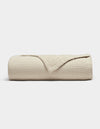 The Cozy Earth Cotton Gauze Blanket, in beige with a soft grid texture, is neatly rolled with a corner folded back to reveal its fabric, shown against a plain white background.|Color:Coconut