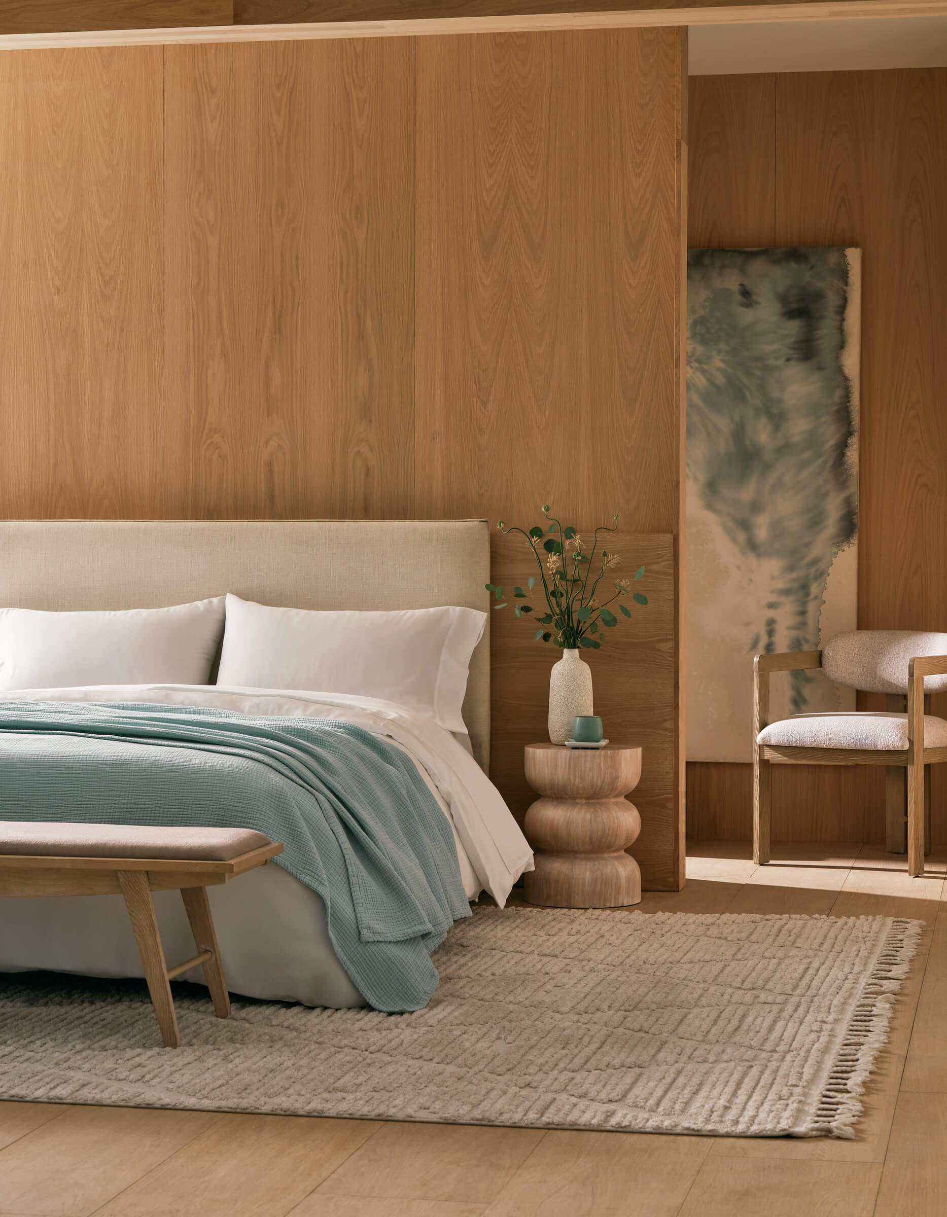A modern bedroom with light wood-paneled walls, a cream bed topped with Cozy Earth's Cotton Gauze Blanket, white and light blue bedding, a layered side table with greenery, a beige bench, and a soft woven rug.