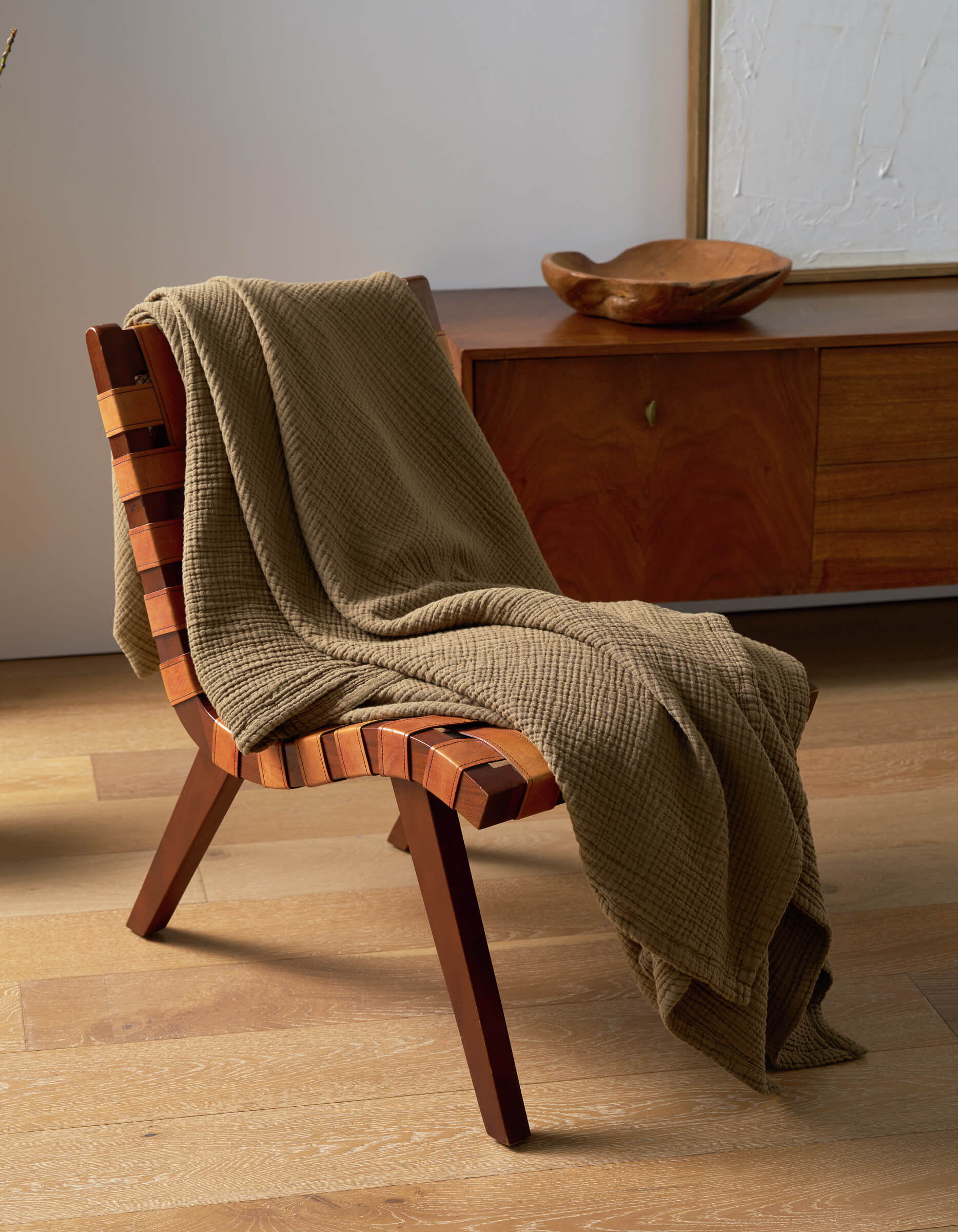 A modern wooden chair with a checkered seat rests on a light wood floor. Draped over it is the Cozy Earth Cotton Gauze Blanket in textured olive-green. In the background, a wooden sideboard and framed artwork are visible.