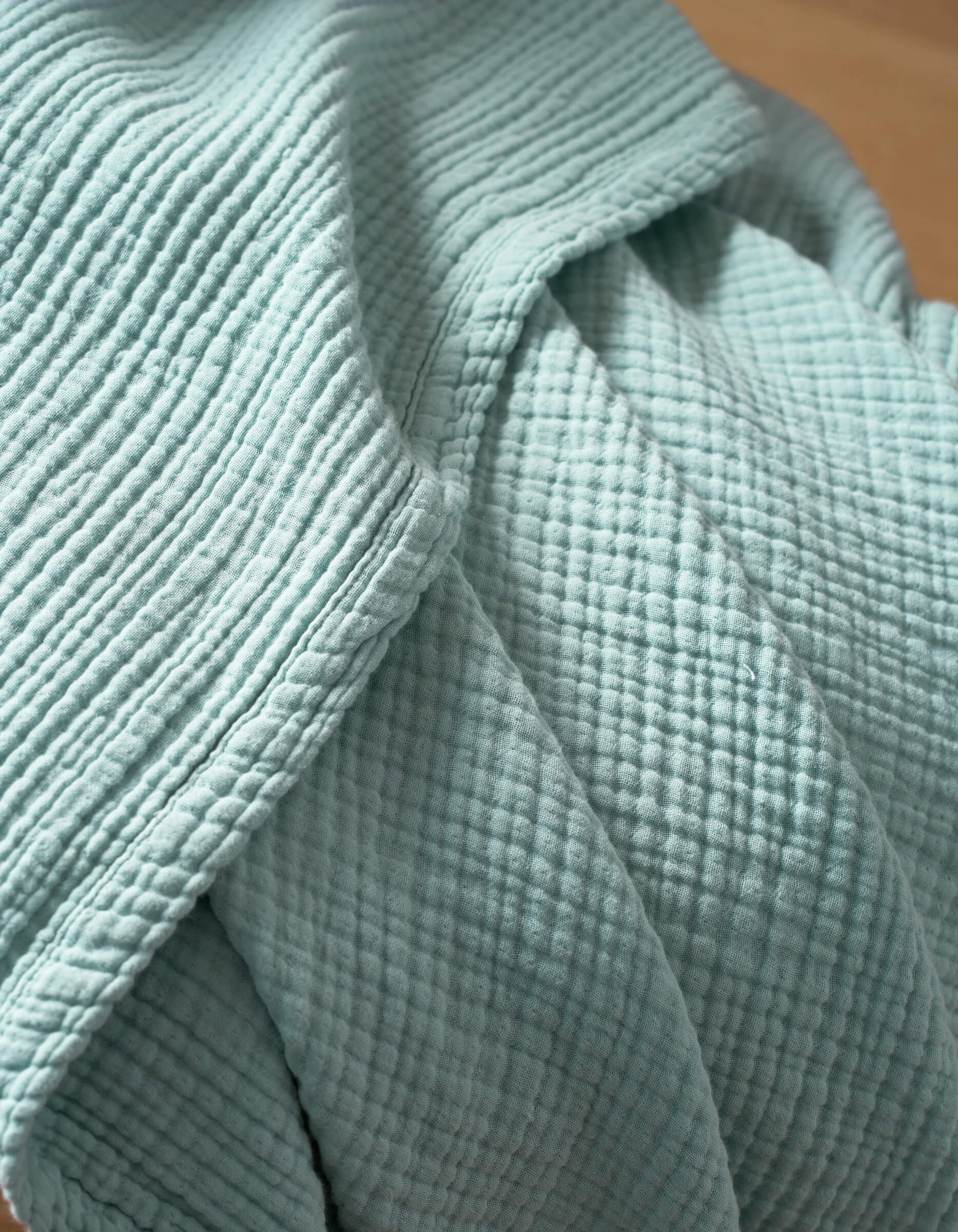 A close-up of Cozy Earth's Cotton Gauze Blanket highlights its textured, light blue woven fabric with a grid pattern, soft folds, and stitched seams.
