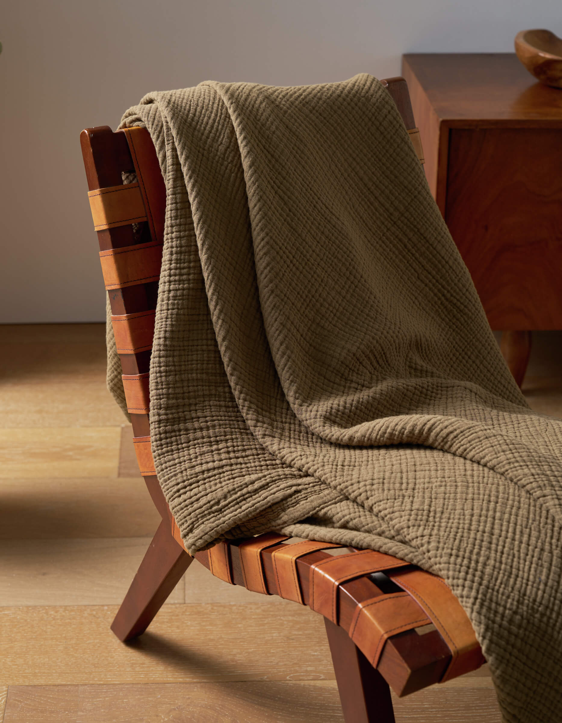 A Cozy Earth Cotton Gauze Blanket in olive green is draped over a wooden chair with a woven leather seat, set on a light wood floor beside another wooden furniture piece.