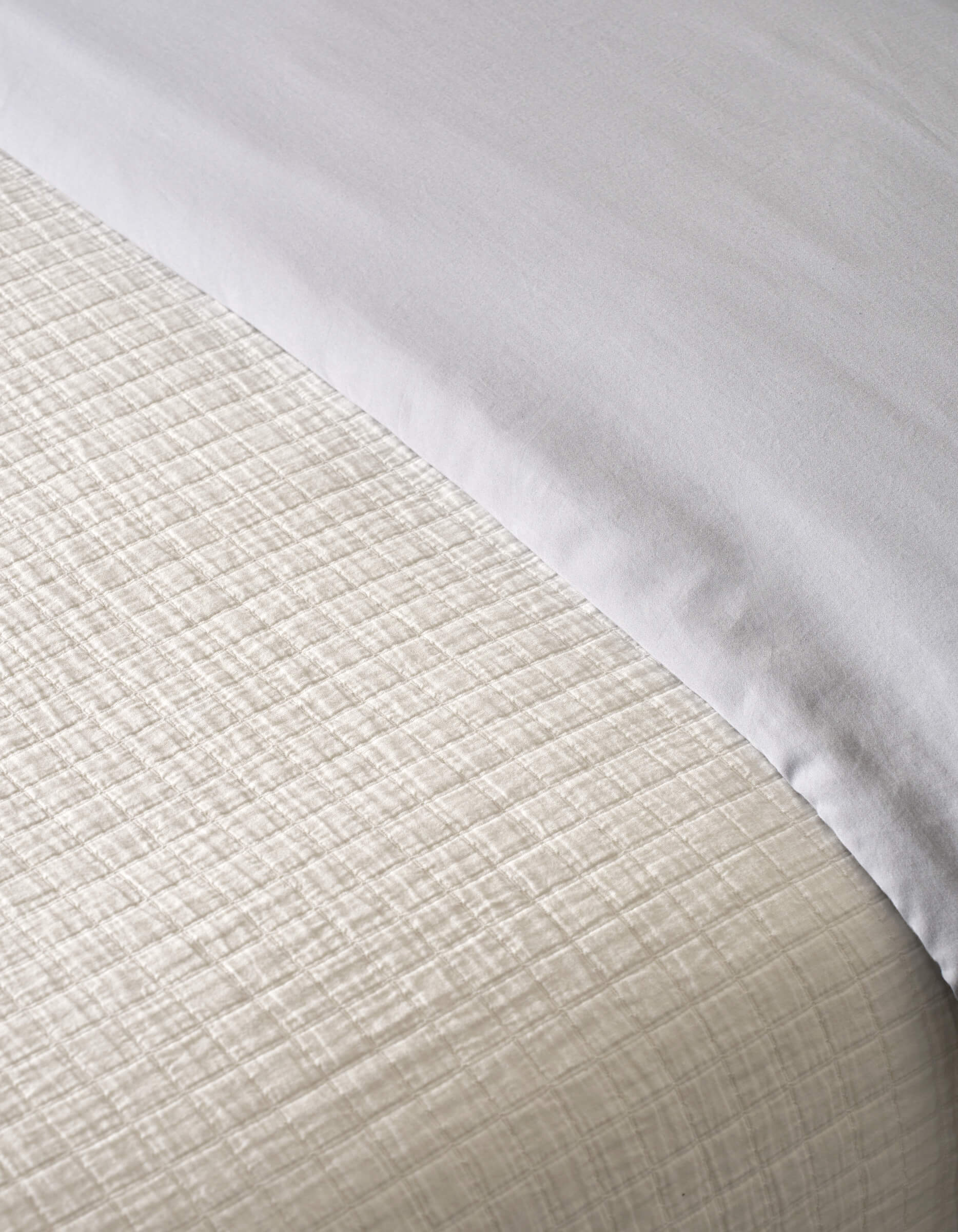 A close-up view shows a neatly made bed featuring the Cozy Earth Waterfront Grid Matelasse Blanket, its textured cream fabric with a subtle grid pattern beneath a smooth white top sheet folded over at the edge.