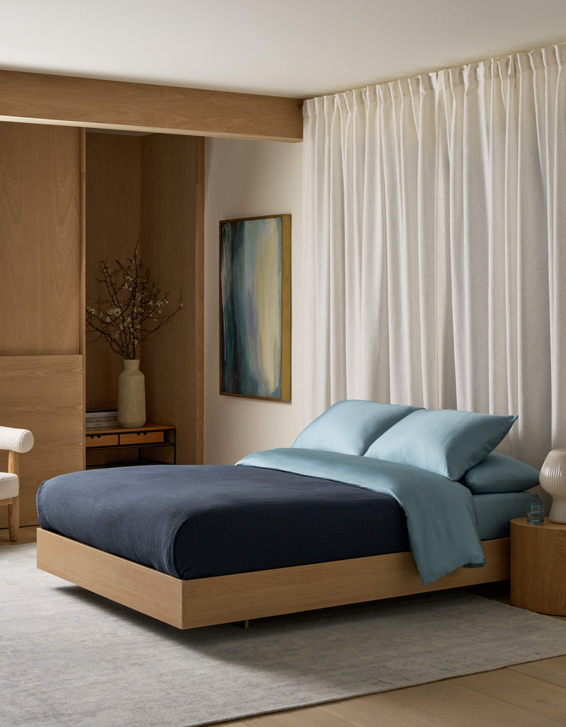 A modern bedroom features a light wood bed frame, the Waterfront Grid Matelasse Blanket by Cozy Earth in navy, light blue pillows, a large abstract painting, and a vase with branches on a wooden desk near floor-length white curtains.