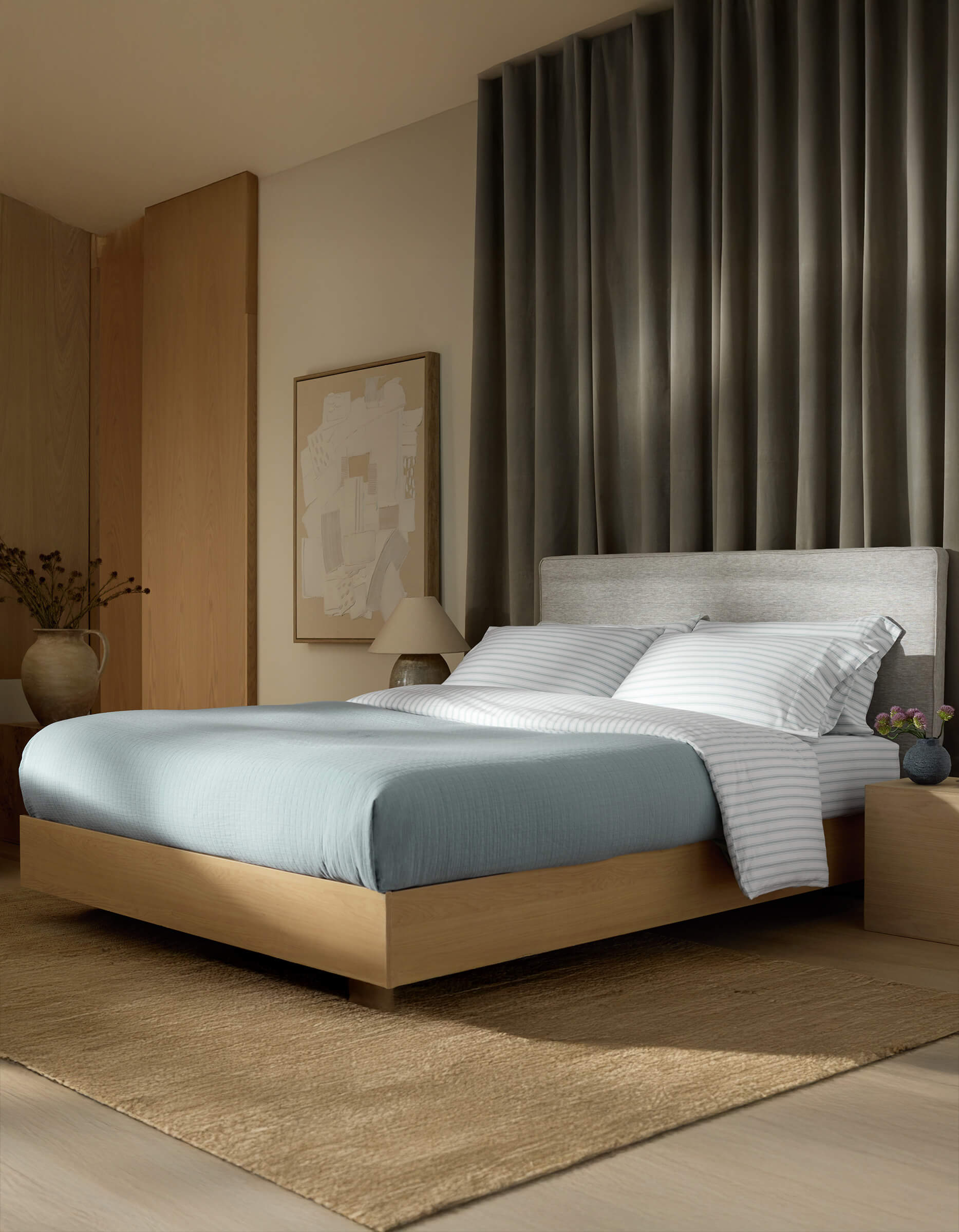 A modern bedroom features Cozy Earth’s Waterfront Grid Matelasse Blanket in blue, a light wood platform bed, striped white pillows, gray upholstered headboard, neutral rug, nightstand with vase, large abstract wall art, and dark curtains behind the bed.
